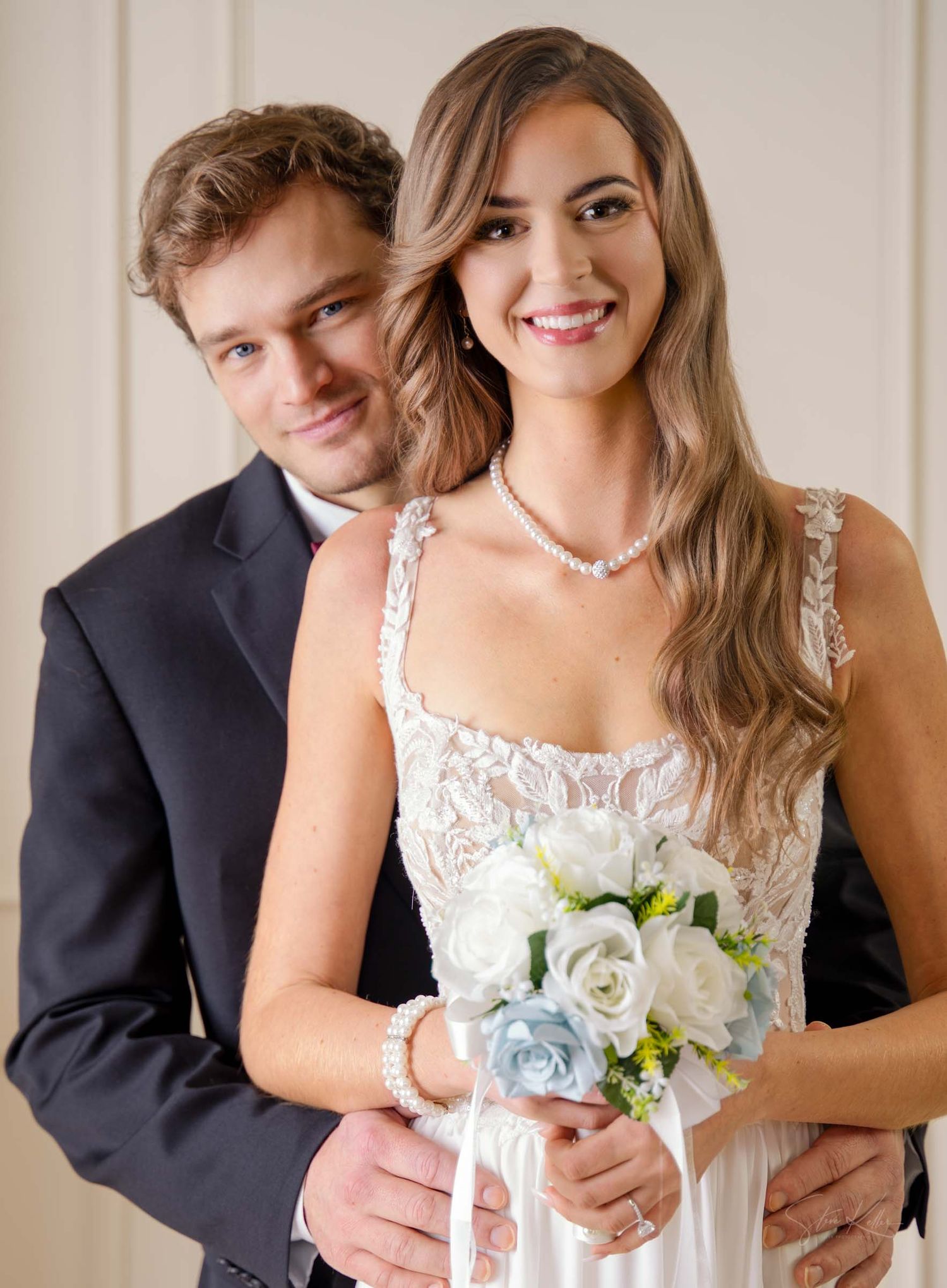 A couple in wedding attire poses together, with the bride holding a bouquet of white and light blue roses.