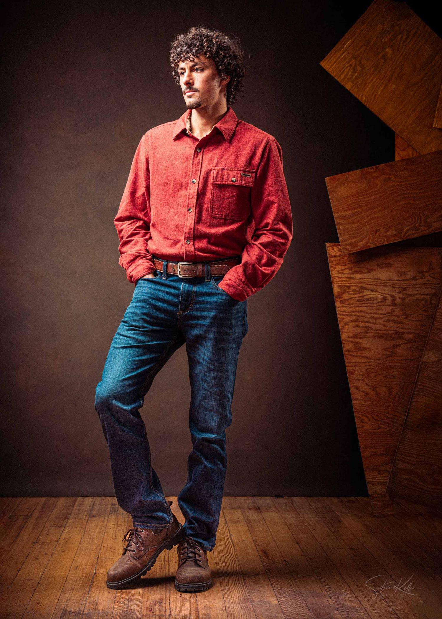 Person in a red button-down shirt and blue jeans poses against a dark wooden backdrop in a casual studio setting.
