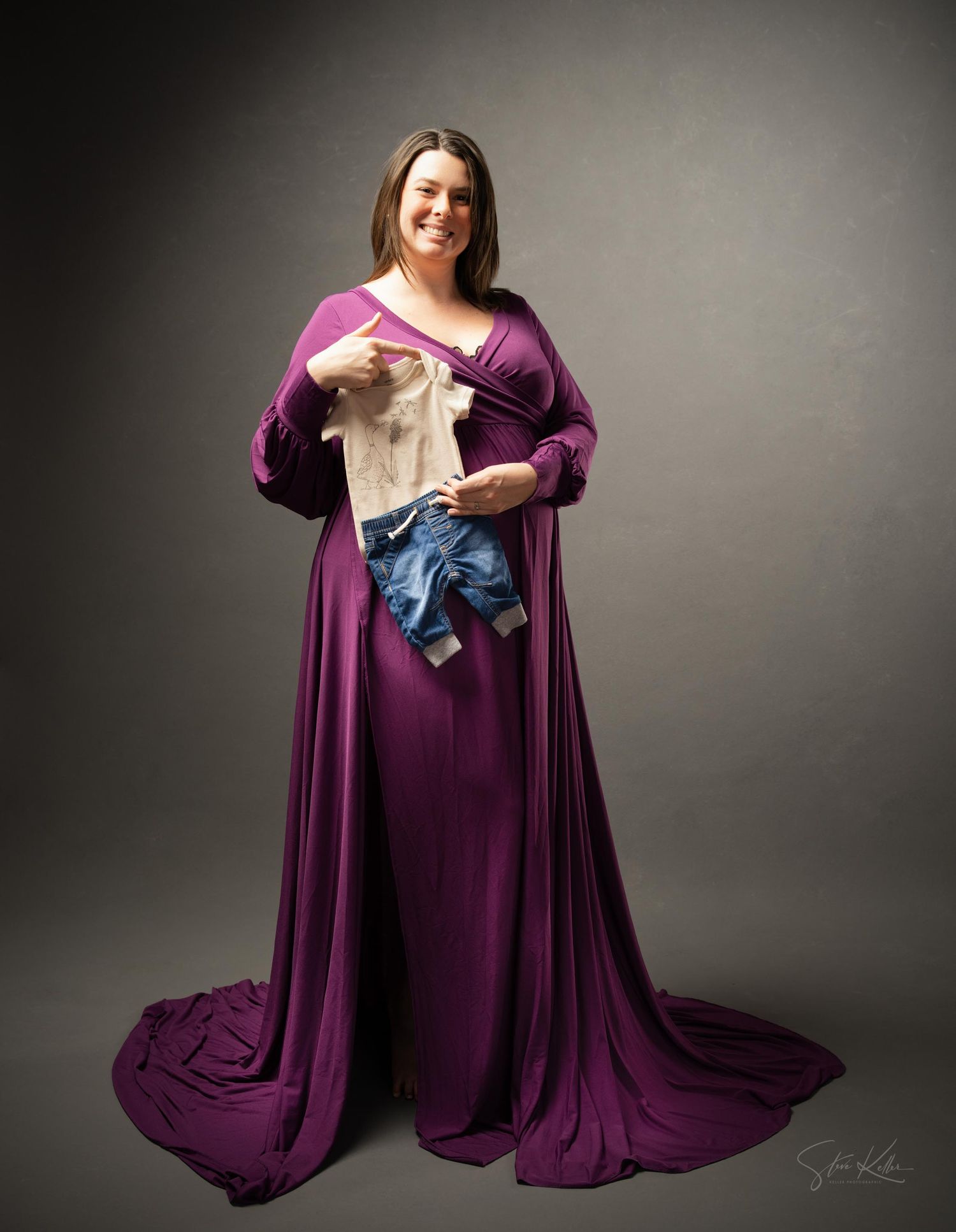 Person wearing a flowing purple maternity gown holds baby clothes against grey studio background.