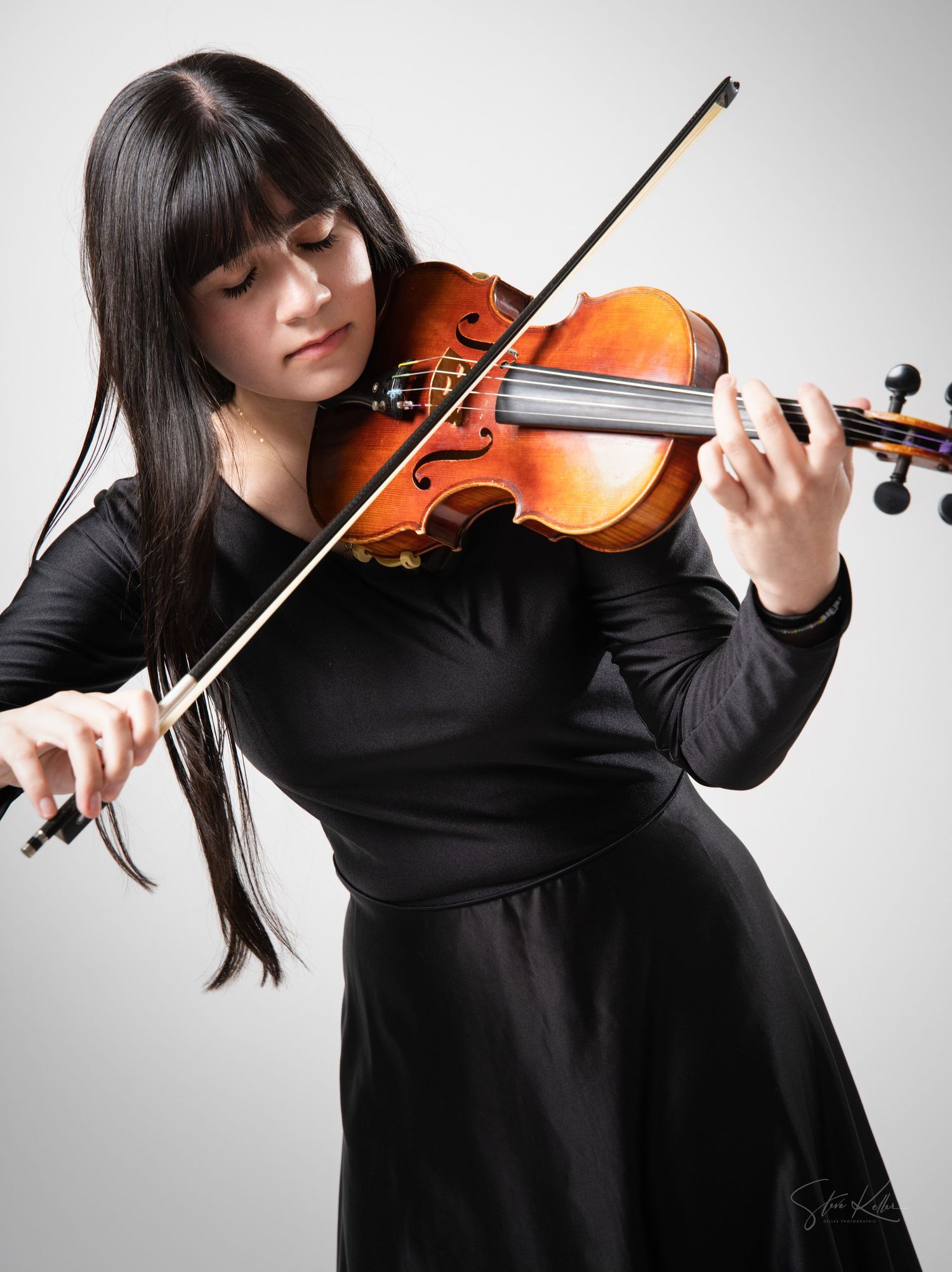 Musician in long black dress plays classical violin while performing against light gray background.
