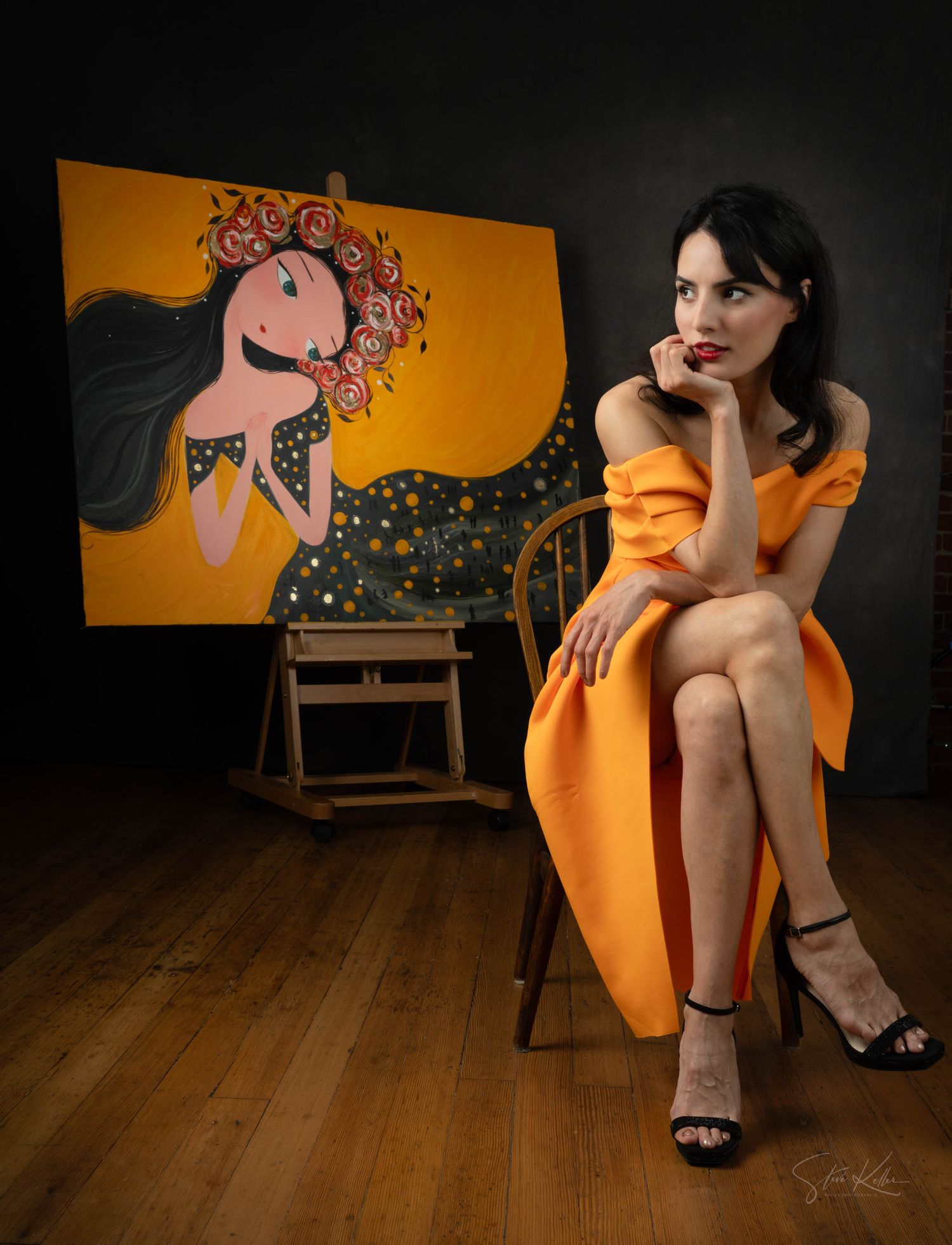 A figure in an orange dress sits thoughtfully in a dark room with a whimsical portrait painting on the orange wall behind.