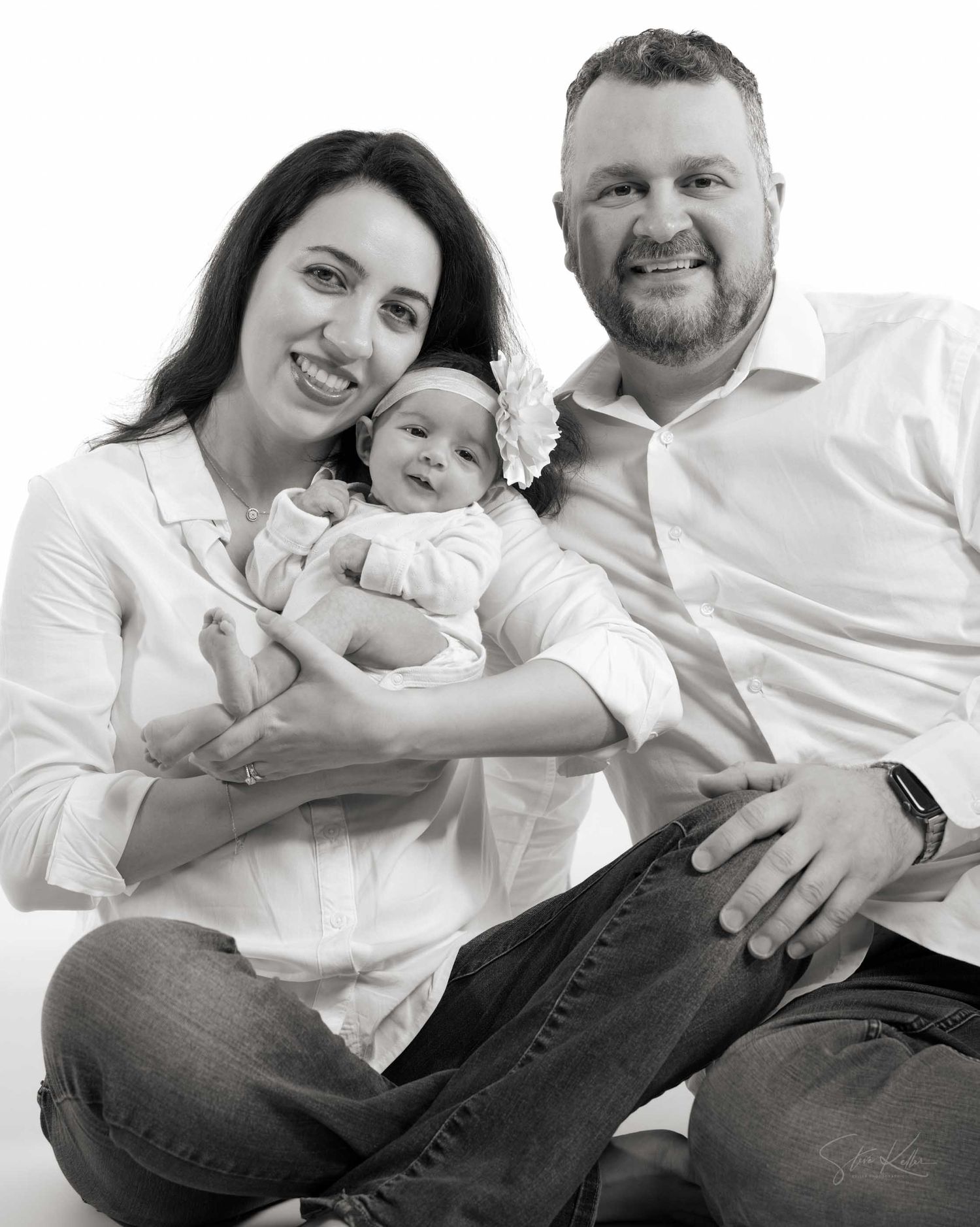 Black and white family portrait of smiling parents holding their newborn baby in white outfits.