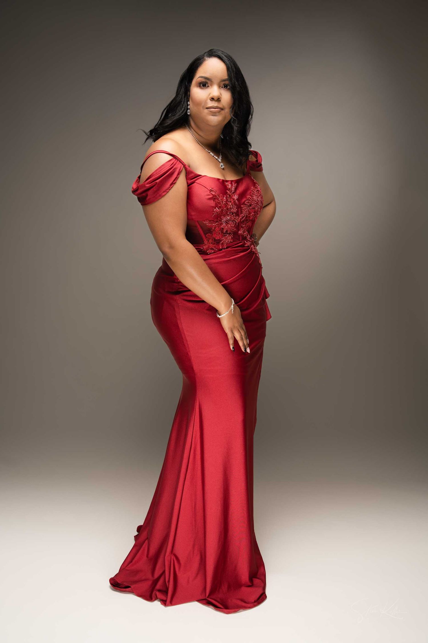 Woman in elegant red off-shoulder mermaid gown with floral embroidery posing gracefully in a studio setting.