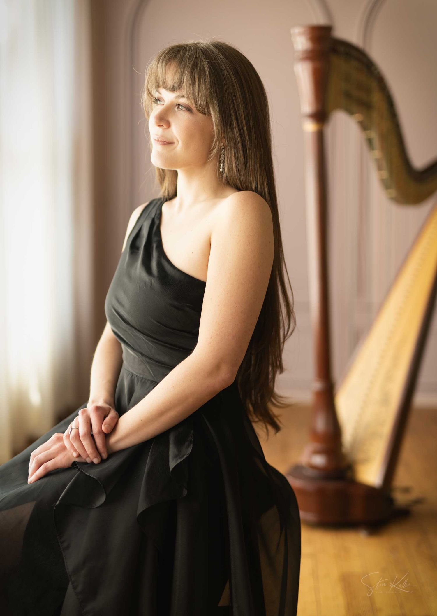 A female harpist in a black gown sits gracefully beside a wooden harp in a softly lit room.