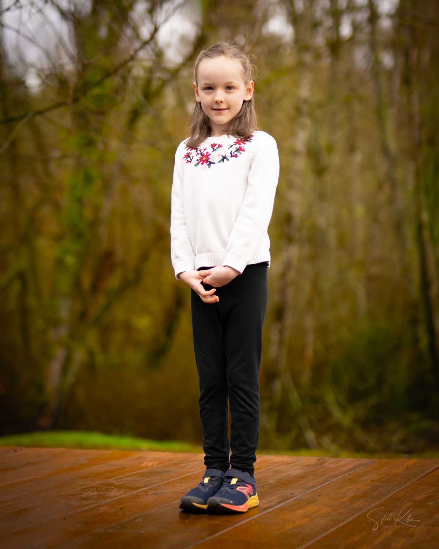 A small child wearing a black and white outfit stands on a wooden platform in a natural outdoor setting.