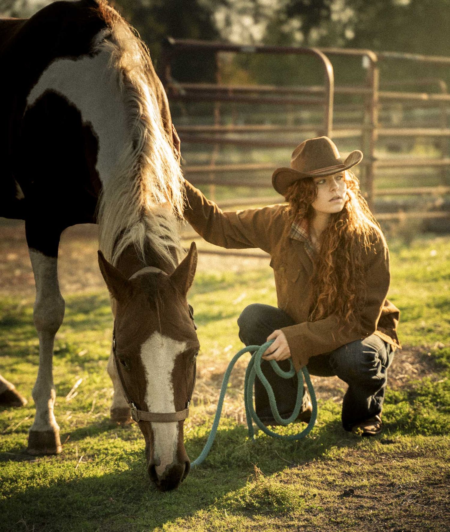 A cowgirl in a brown jacket and hat crouches beside a paint horse on a sunlit ranch, holding a blue lead rope.