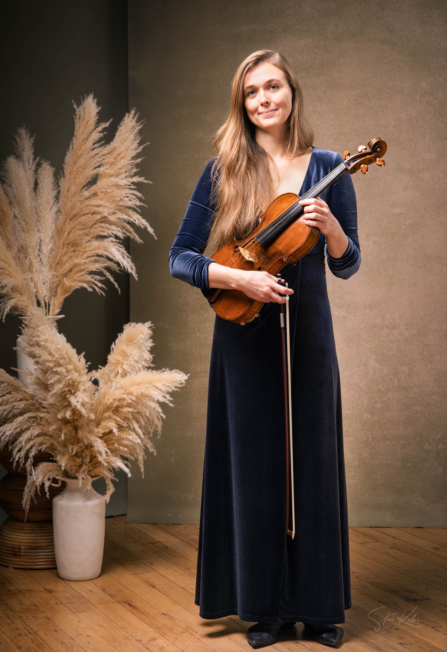 A violinist in a navy blue dress performs next to a vase of pampas grass in a studio setting.