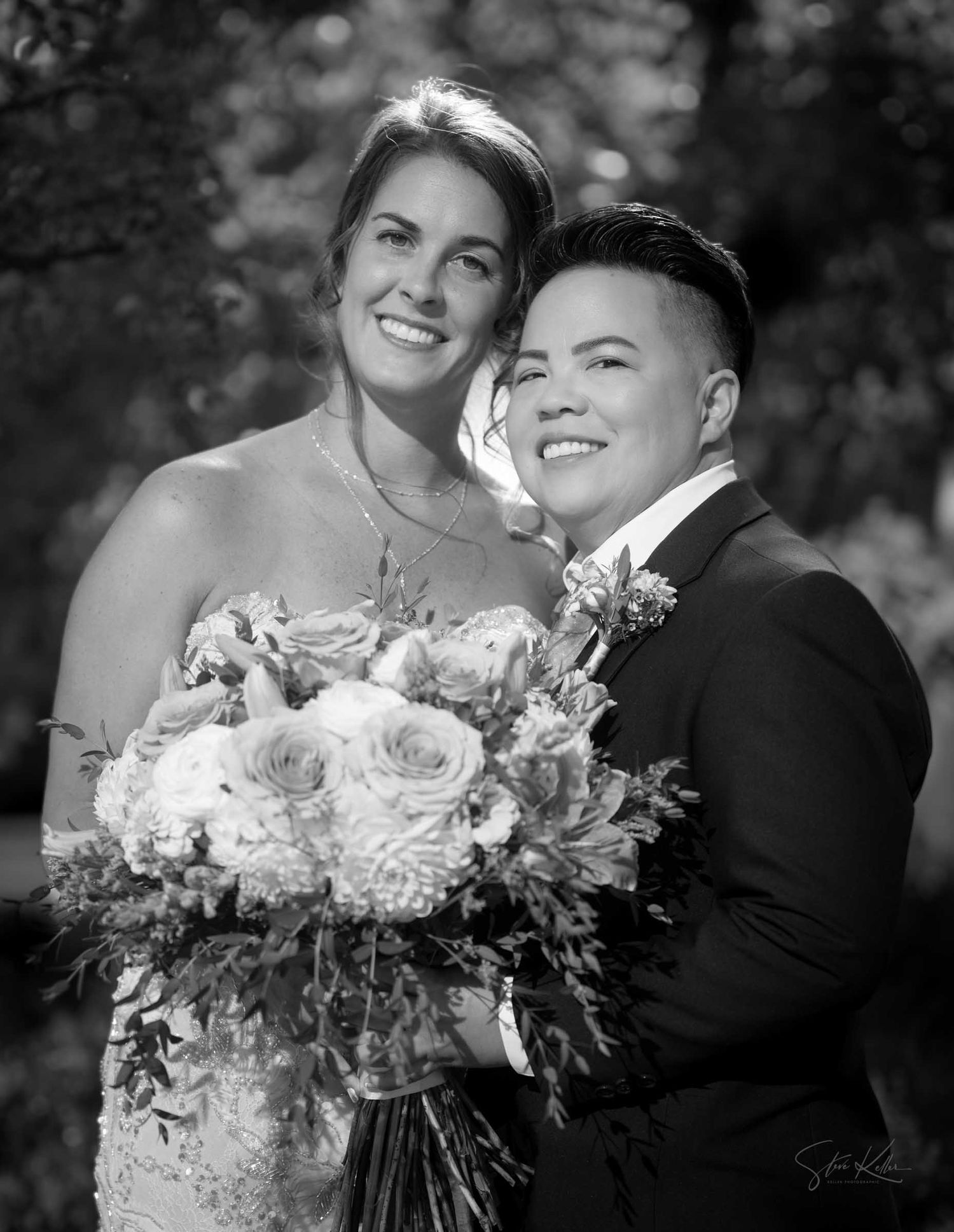 Black and white portrait of smiling bride and partner in tuxedo holding floral bouquet outdoors.