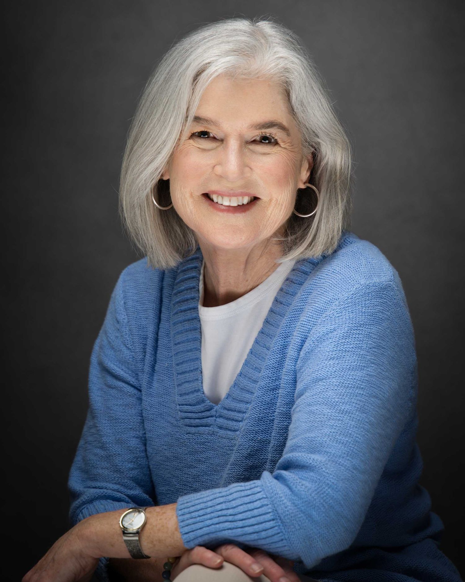 Smiling older woman with gray hair wearing a blue cardigan sweater and hoop earrings poses for a professional portrait.