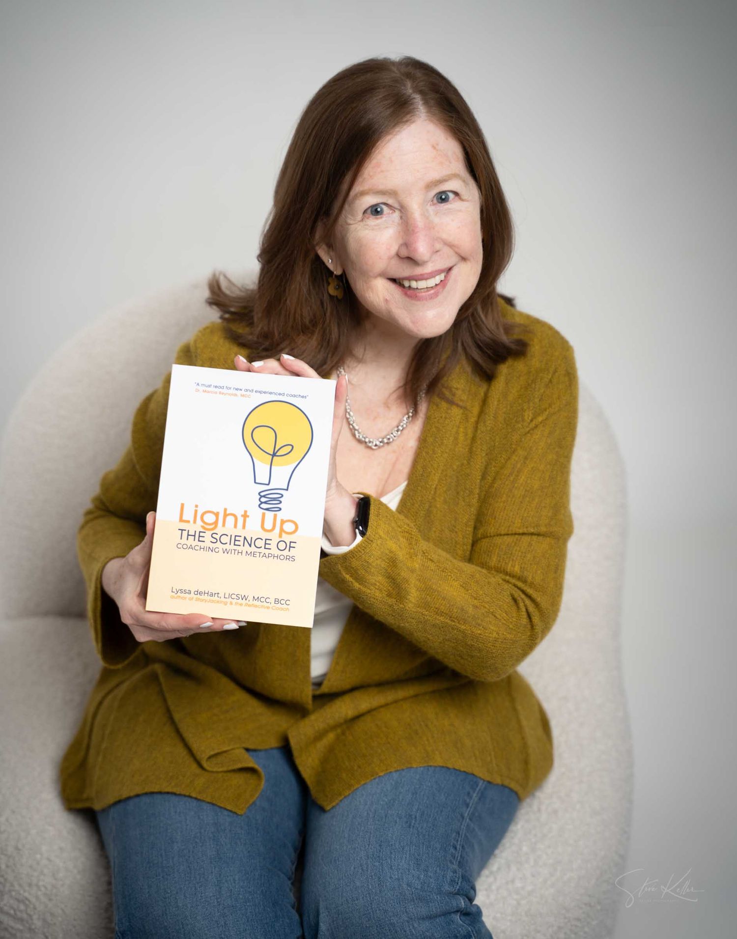 Smiling woman in mustard cardigan holding Light Up The Science Of book while seated in white chair.