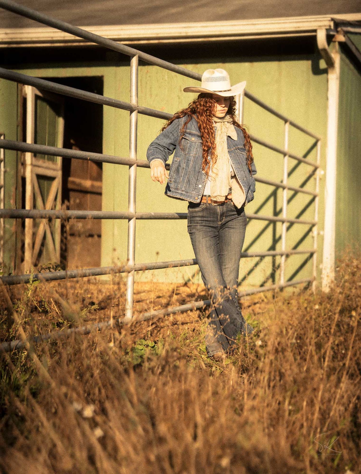Woman in denim jacket, jeans, and white cowboy hat standing by a metal fence on a rustic farm.