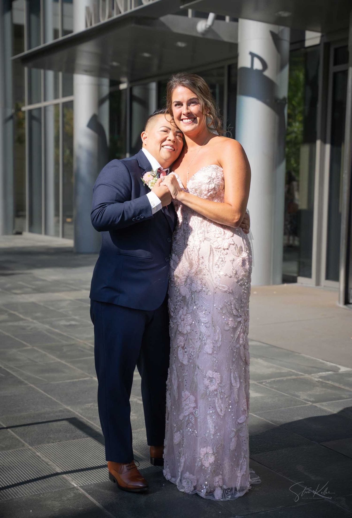 Two brides smile joyfully outside a modern building, one in a navy suit and one in a lace wedding gown.