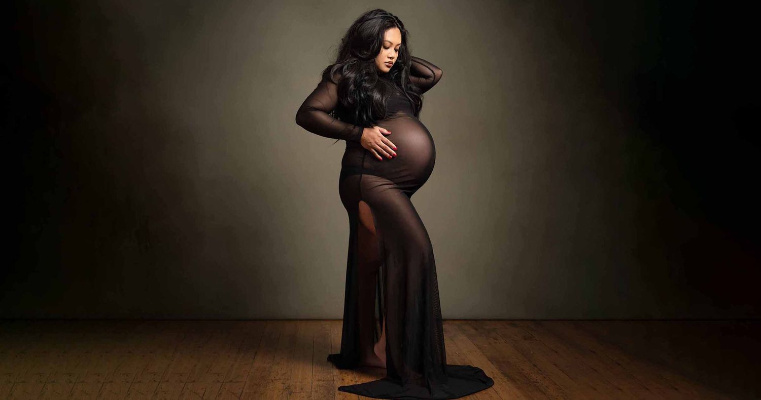 Pregnant woman in a sheer black maternity gown cradles her baby bump during a studio photoshoot against a dark backdrop.
