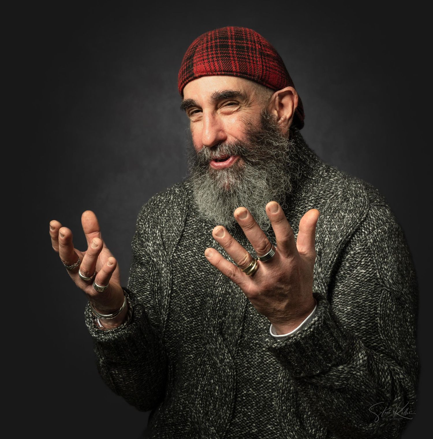 A person in a knit sweater and plaid cap gestures expressively with both hands against a dark background.