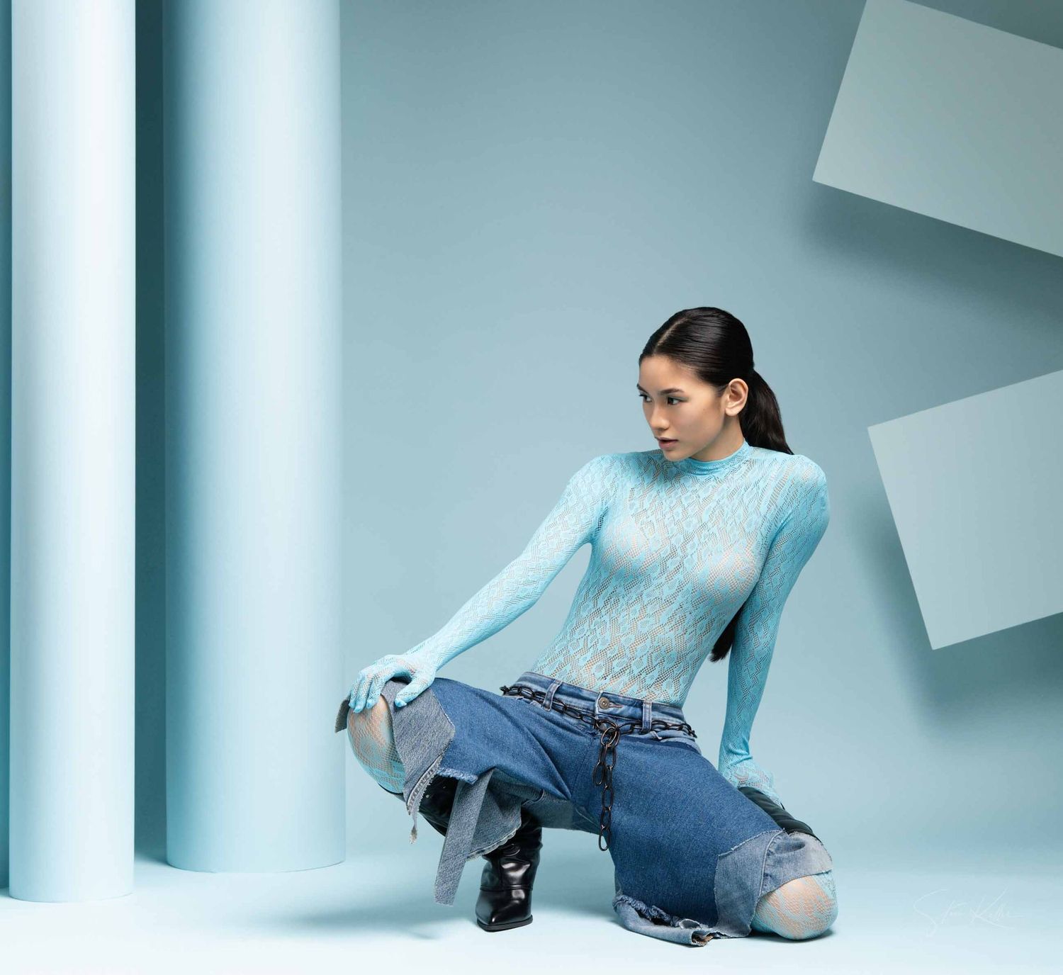 Model in light blue knit sweater and distressed jeans poses on floor against modern geometric backdrop.