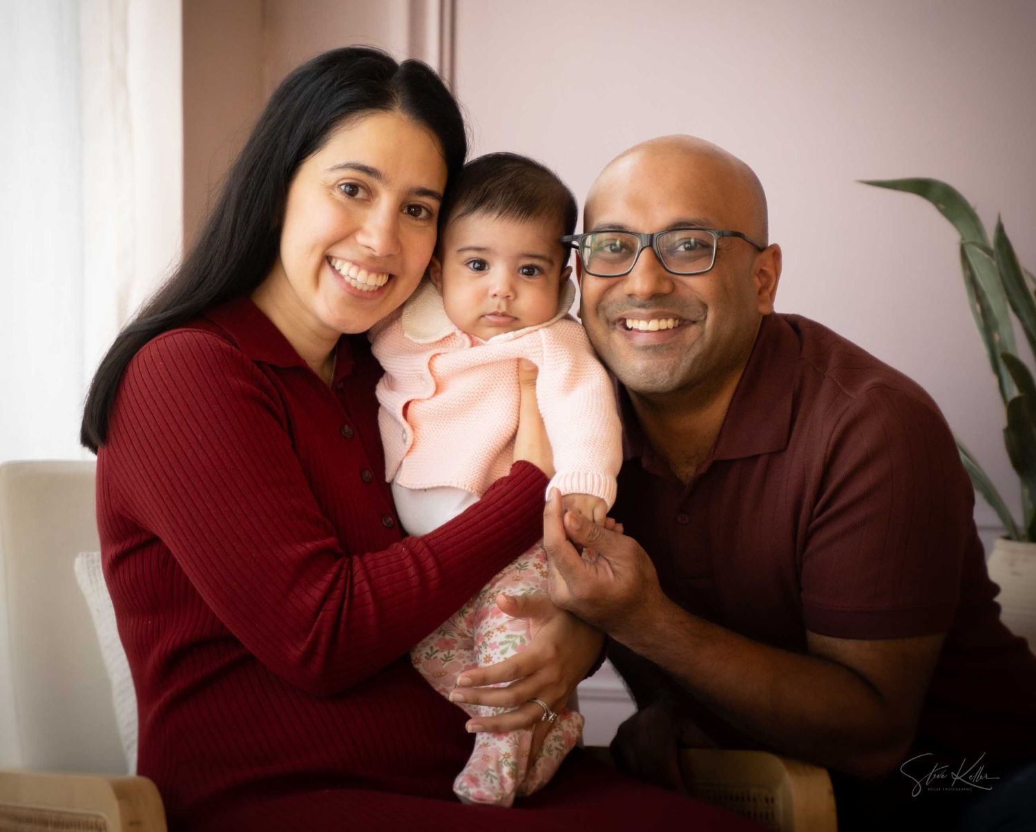 Happy family portrait with smiling parents holding their baby girl in a bright, warmly lit indoor setting.