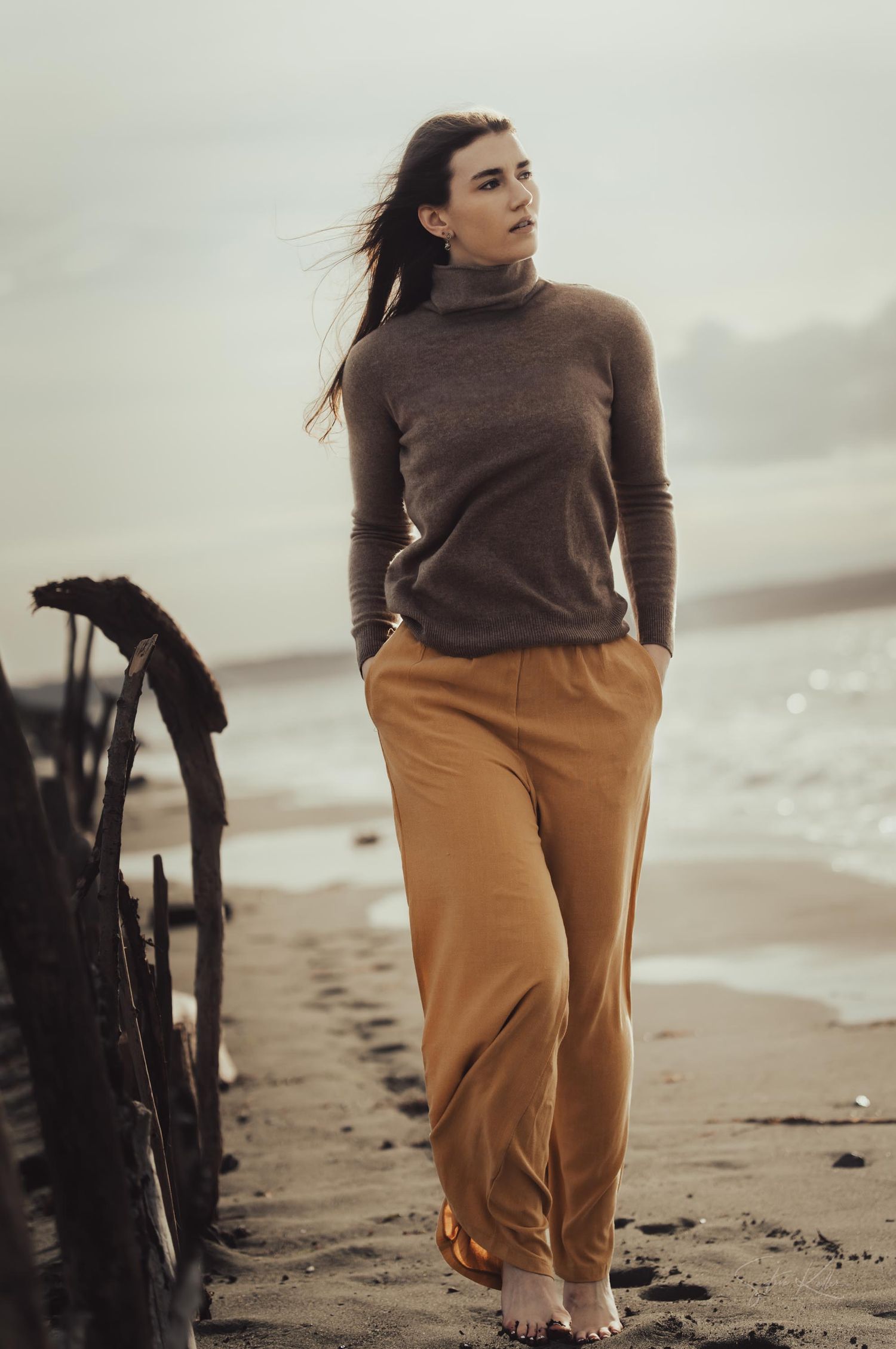 Moody coastal fashion photo with brown turtleneck and tan pants against overcast beach backdrop.