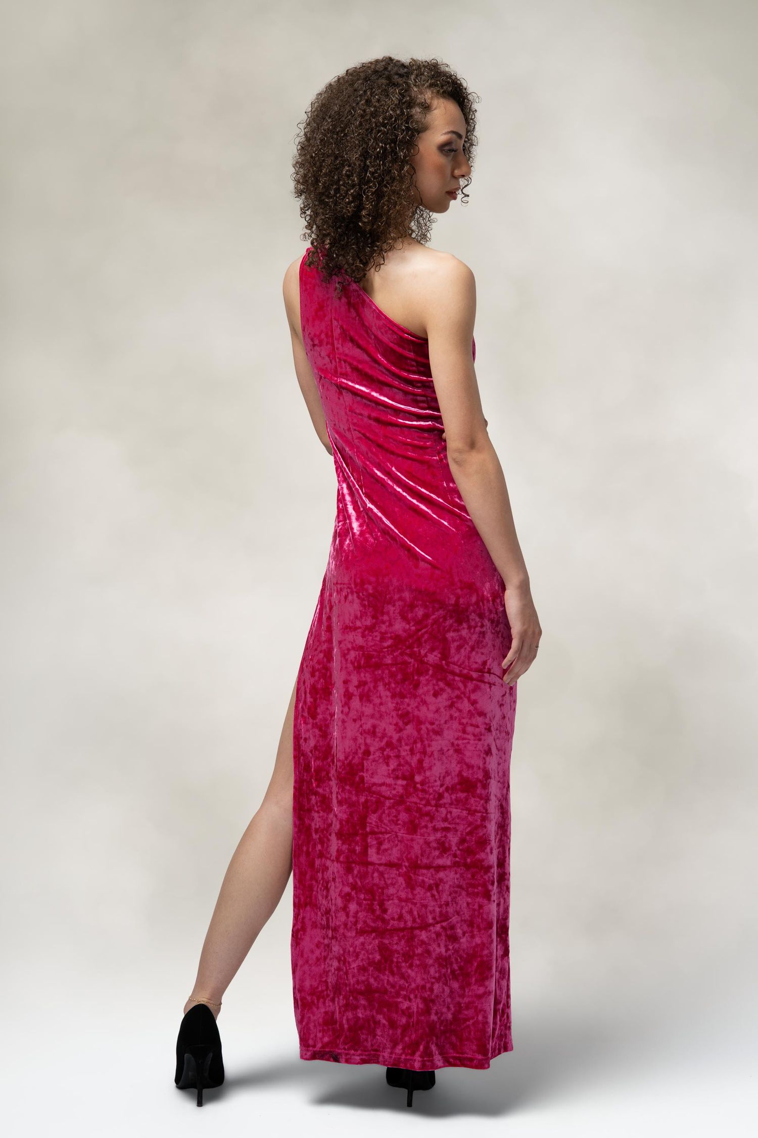 Model poses in elegant red velvet one-shoulder gown with side slit and black heels against neutral background.