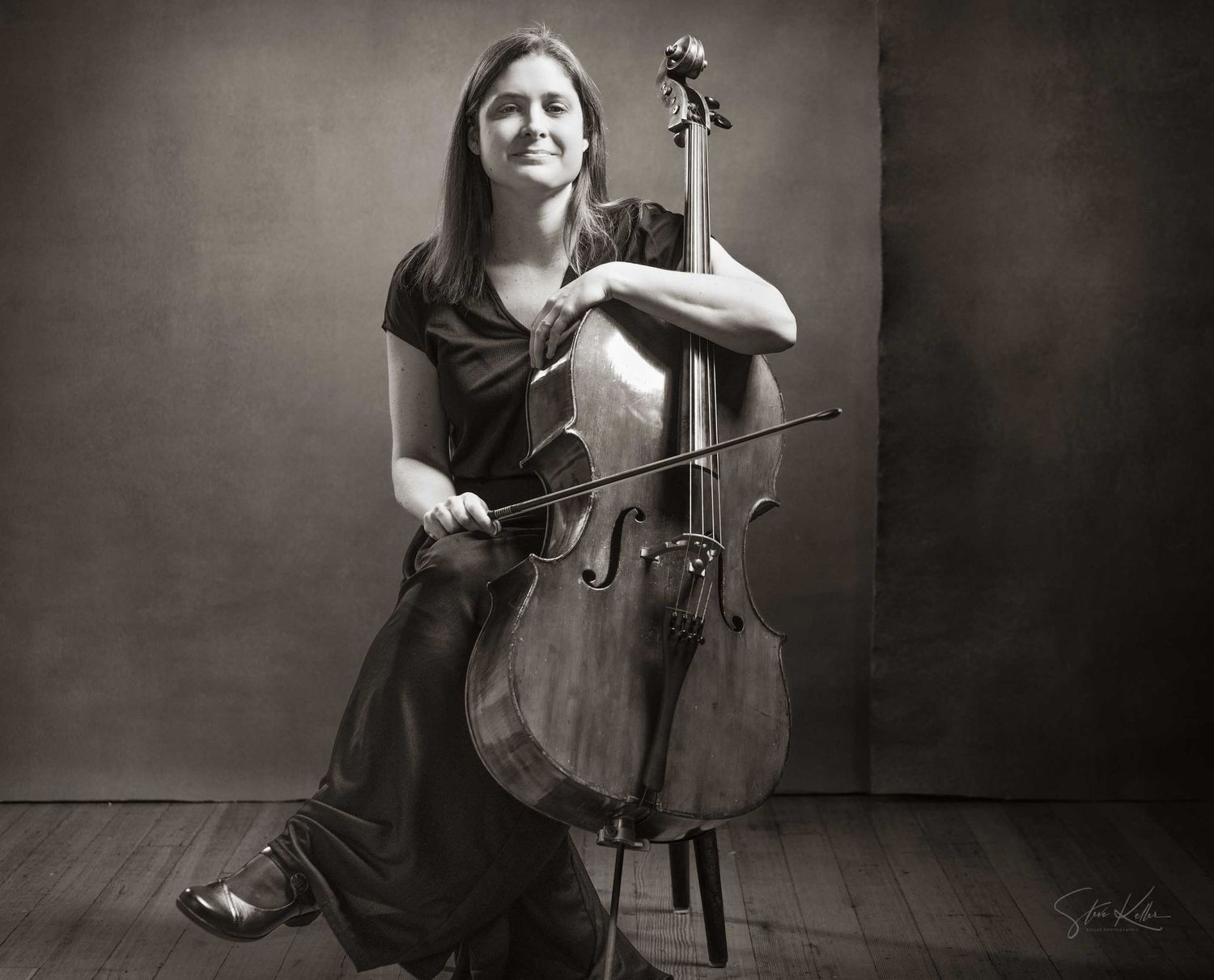Black and white portrait of a smiling woman posing with a cello, holding a bow, seated in a studio setting.