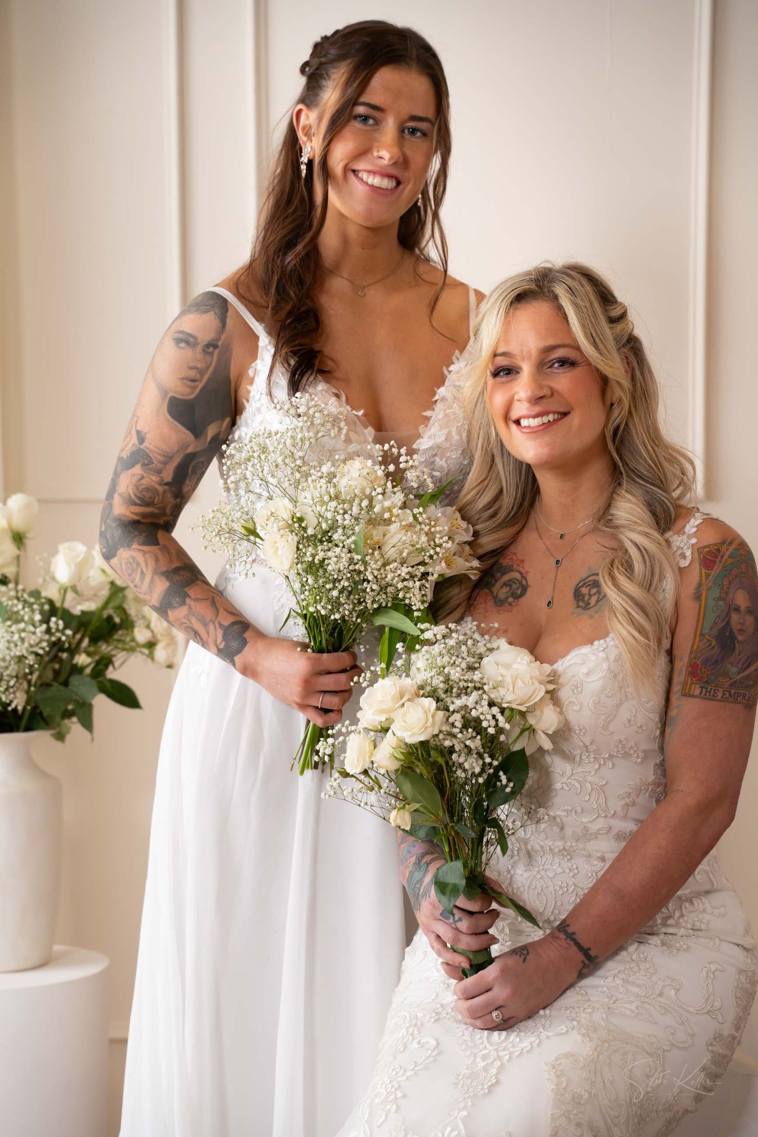 Two brides in white dresses with tattoos pose together holding matching bouquets of white flowers.