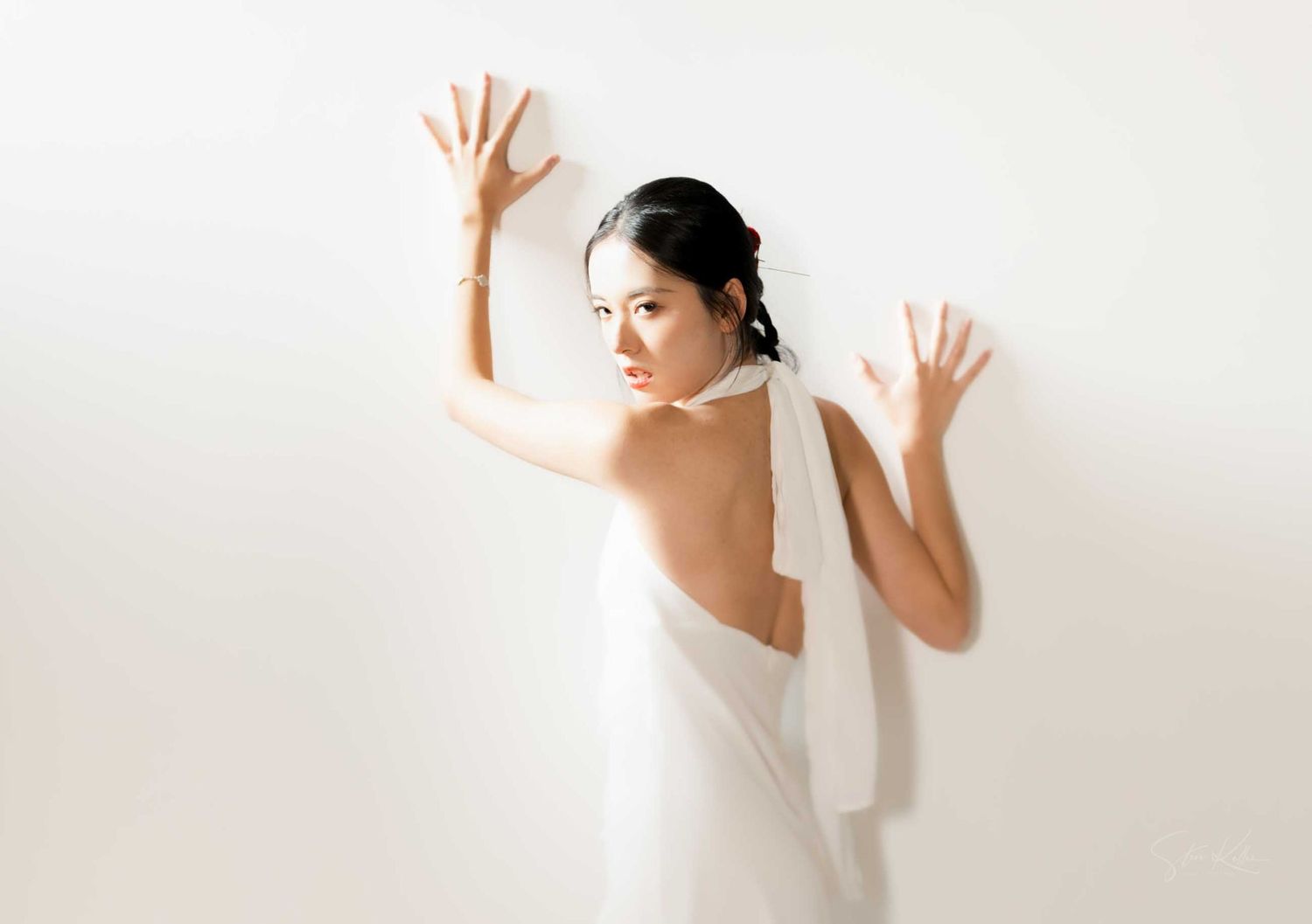 A person in a white dress poses dramatically against a light background with raised hands and an expressive stance.