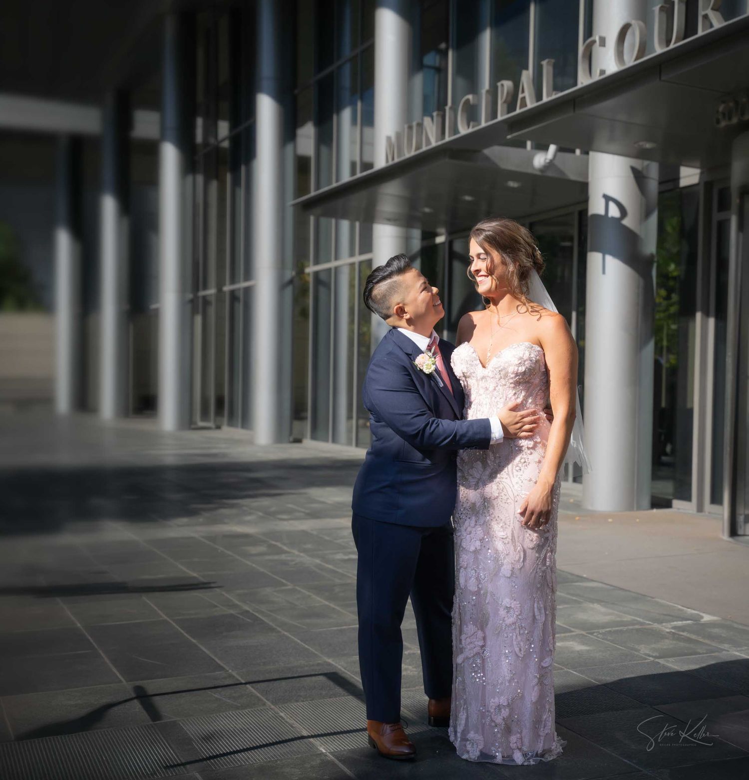 Couple in wedding attire embracing outside a municipal courthouse, bride in lace gown and groom in navy suit.