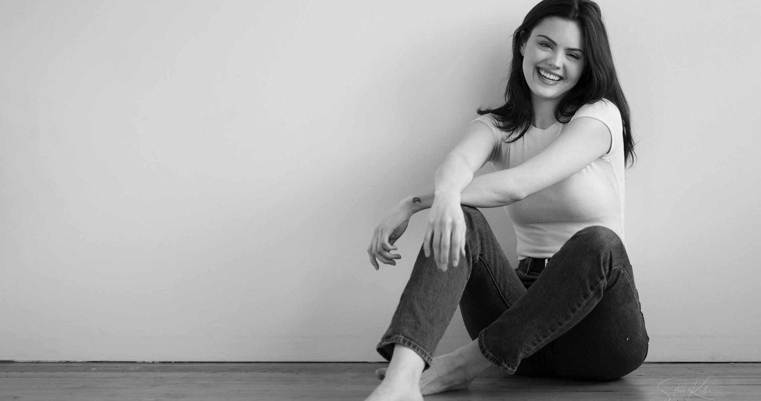 Smiling woman with long dark hair sits barefoot on the floor against a plain wall in jeans and a light top, black and white portrait.