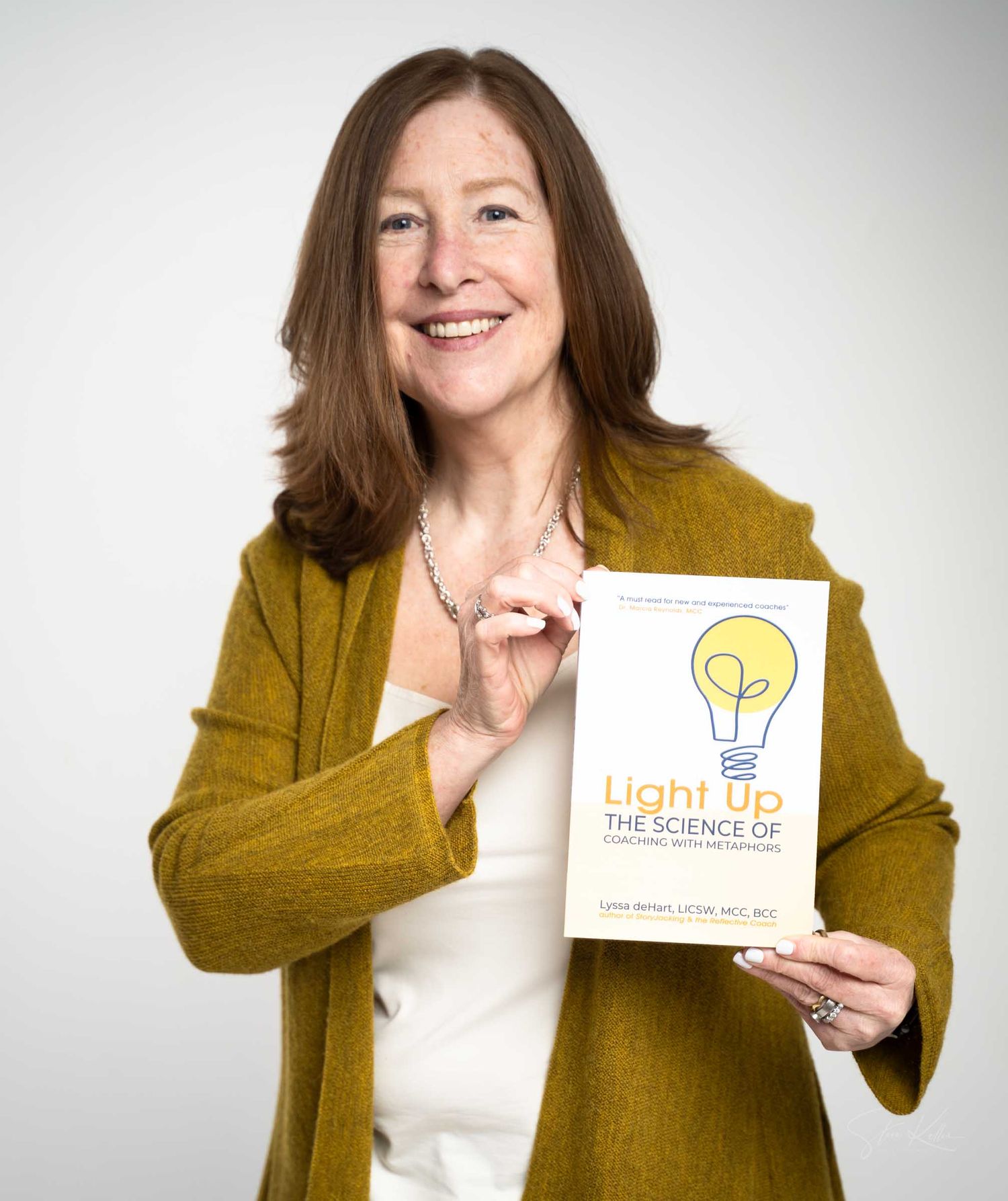 Woman in gold cardigan smiling and holding a book titled Light Up The Science of Learning with Your Brain.