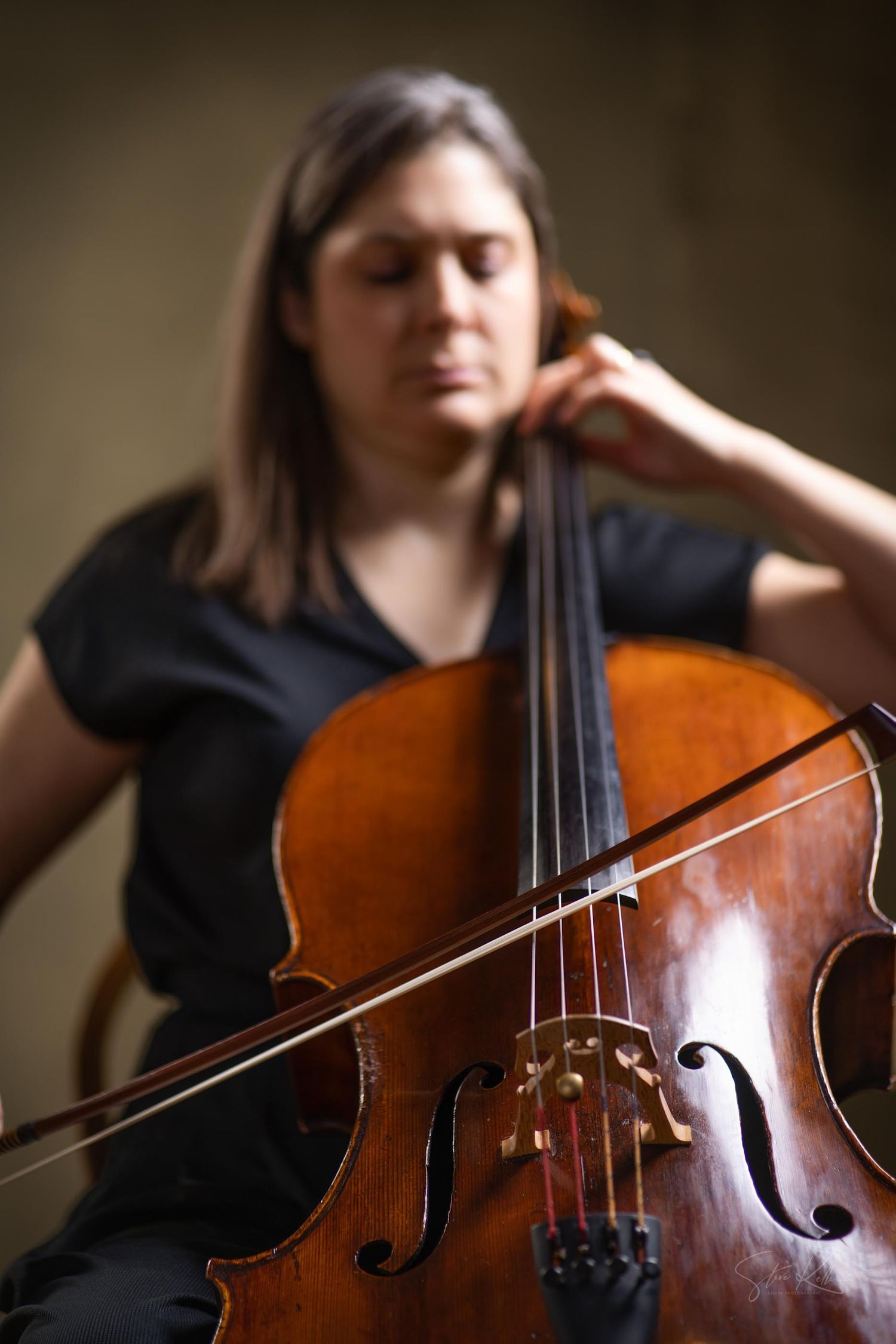 A cellist in a black shirt plays their wooden instrument with a bow in a moody, shallow depth of field portrait.