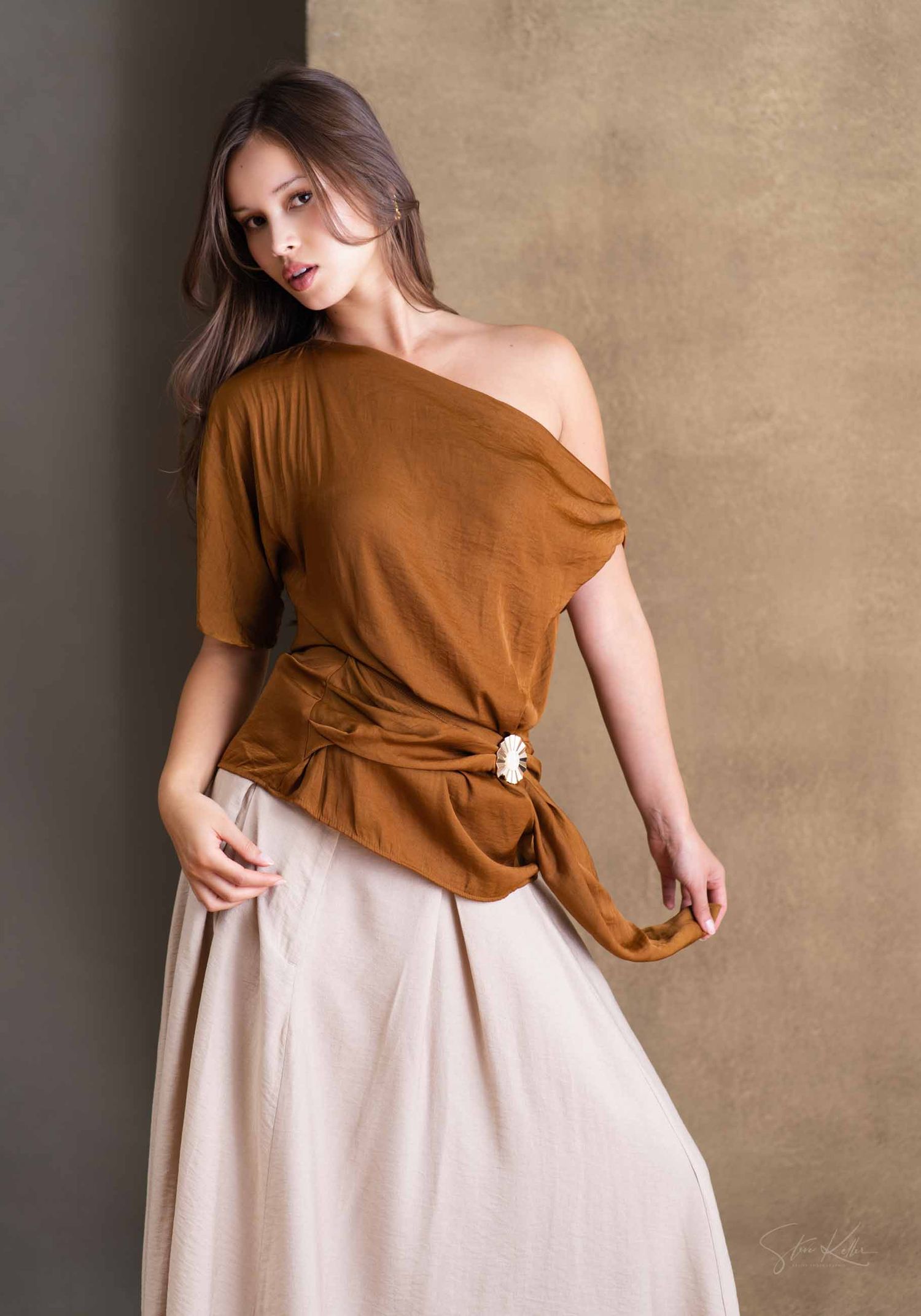 Model in brown asymmetrical top and beige skirt demonstrates various fashion poses against textured wall.