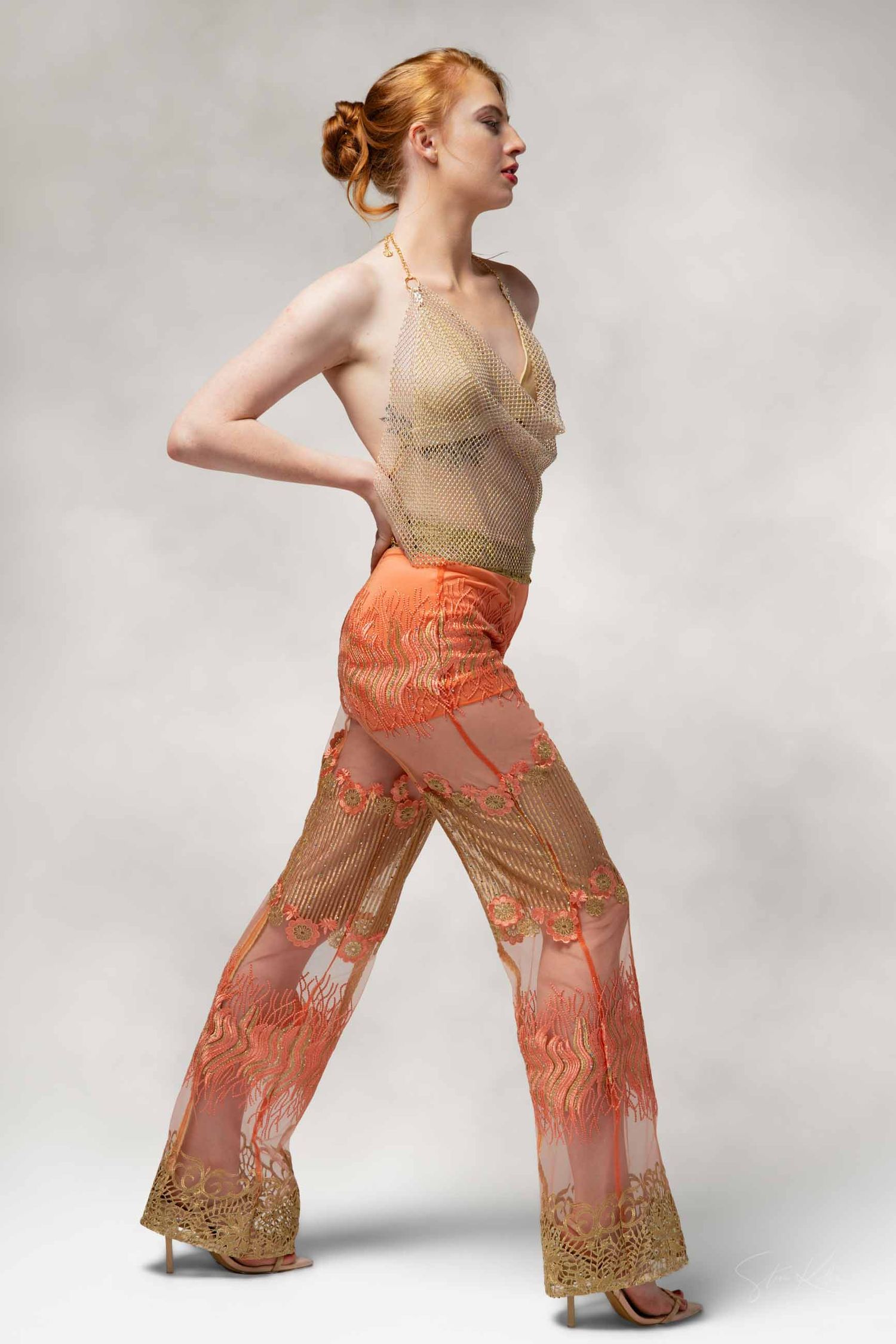 A dancer in a beige and orange lace dress performs an expressive movement sequence against a white backdrop.