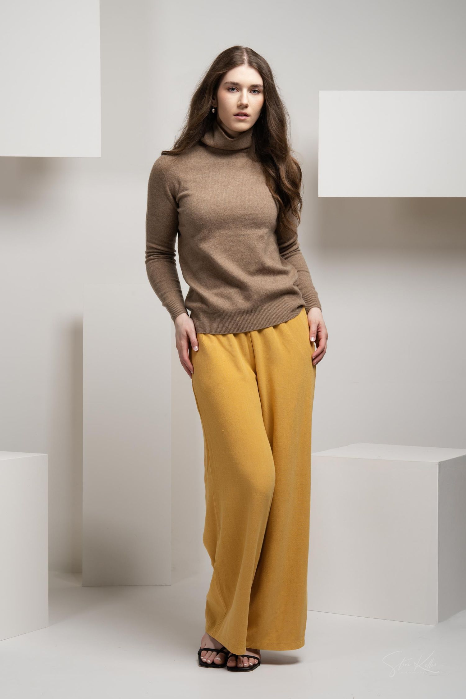 Fashion model wearing brown turtleneck sweater and mustard yellow wide-leg pants against white geometric backdrop.