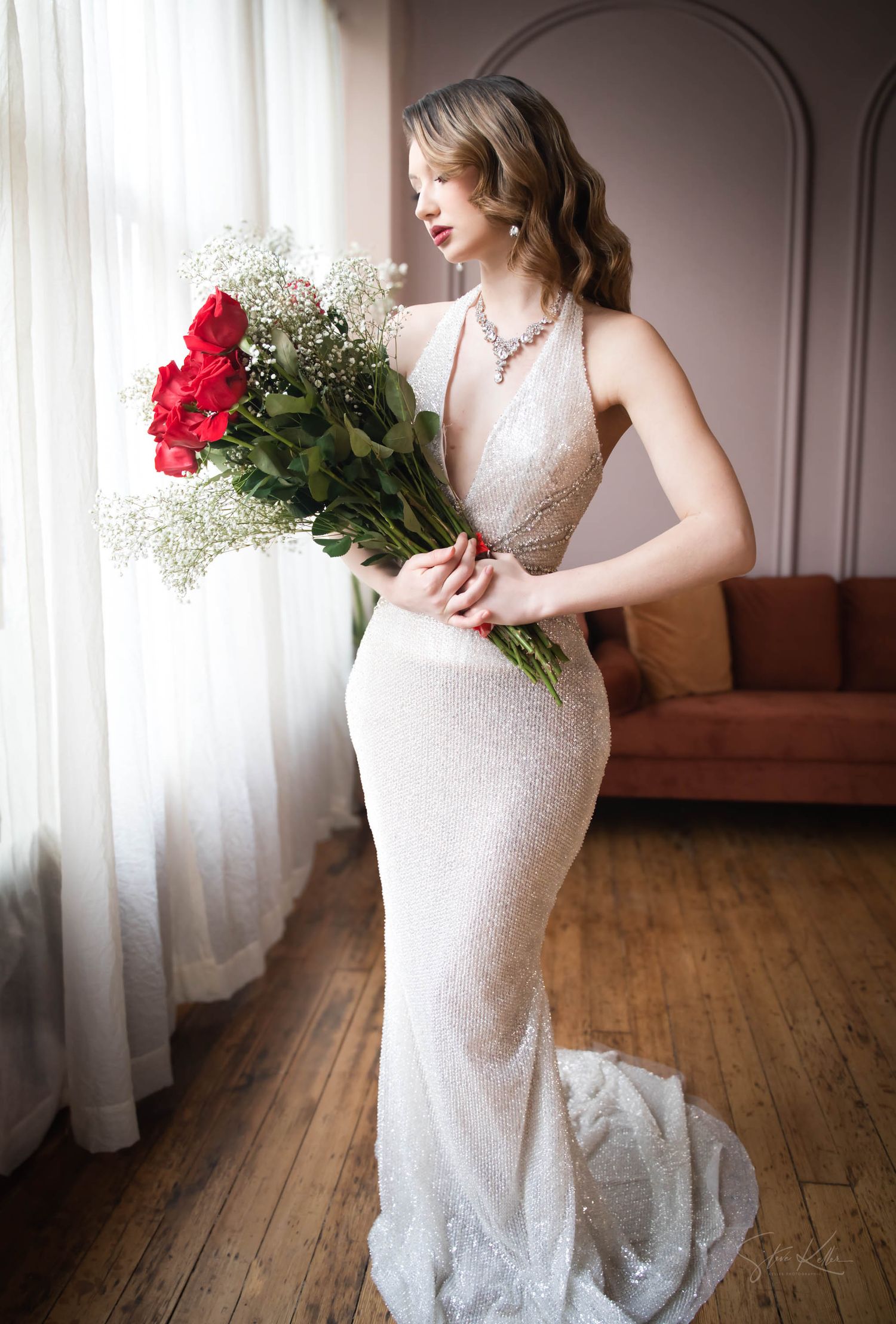 Elegant bride in fitted white lace halter gown holding red roses and baby's breath bouquet by window in sunlit room.
