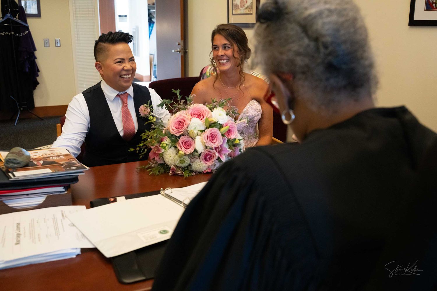 Couple smiling during intimate courthouse wedding ceremony with judge, bride holding pink rose bouquet.