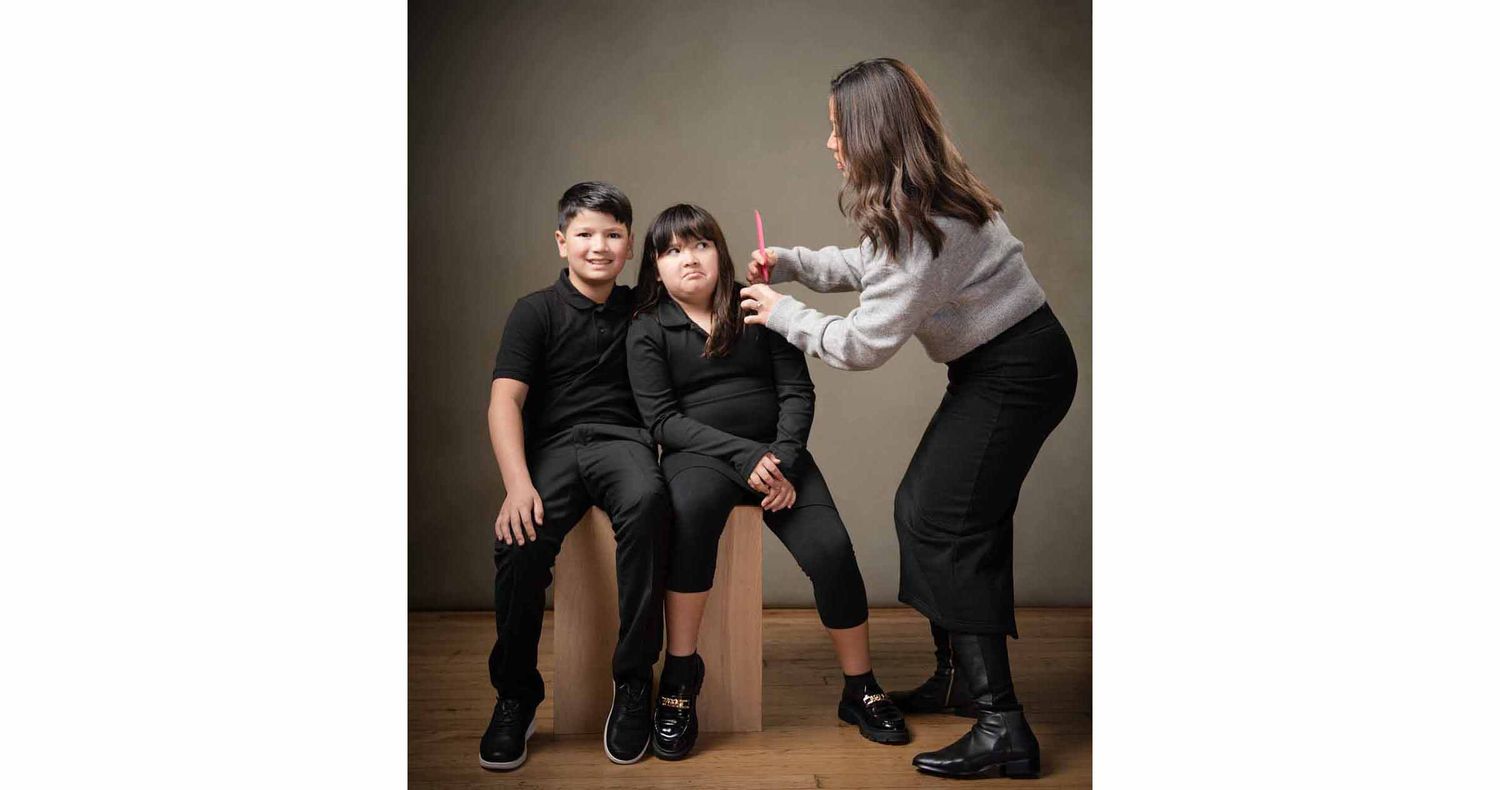 A woman in a gray top adjusts a child's hair while two children in black outfits sit on a wooden stool during a studio portrait session.
