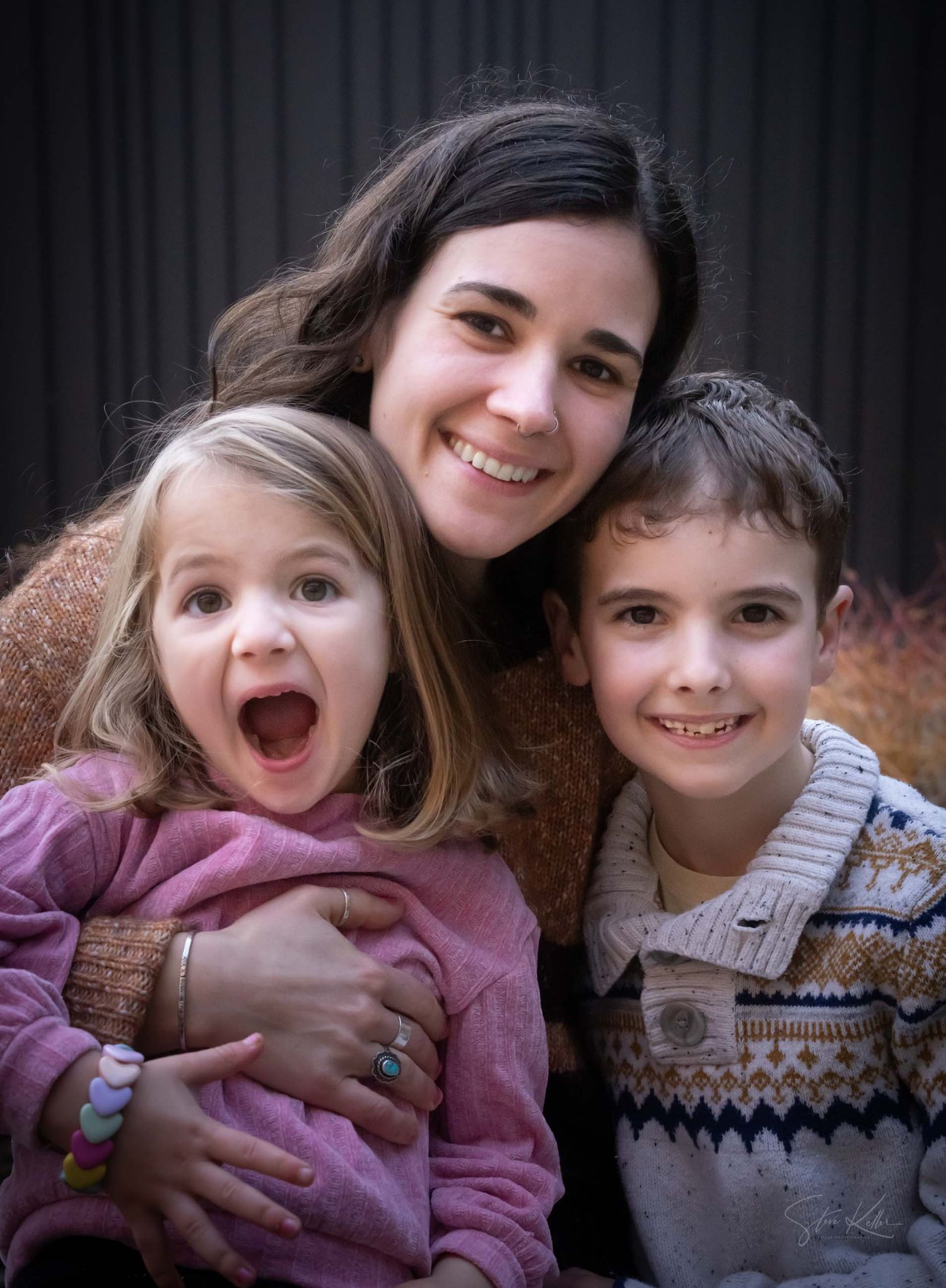 A smiling woman poses with two young children, a girl with an open mouth and a boy, in a warm family portrait.
