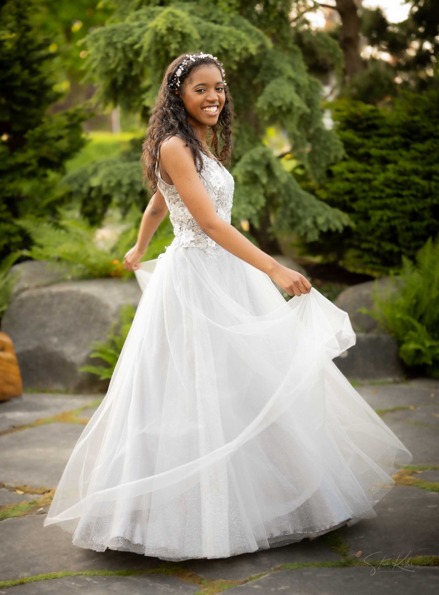 A smiling young woman twirls in a white lace ballgown outdoors, wearing a floral headpiece in a garden setting.