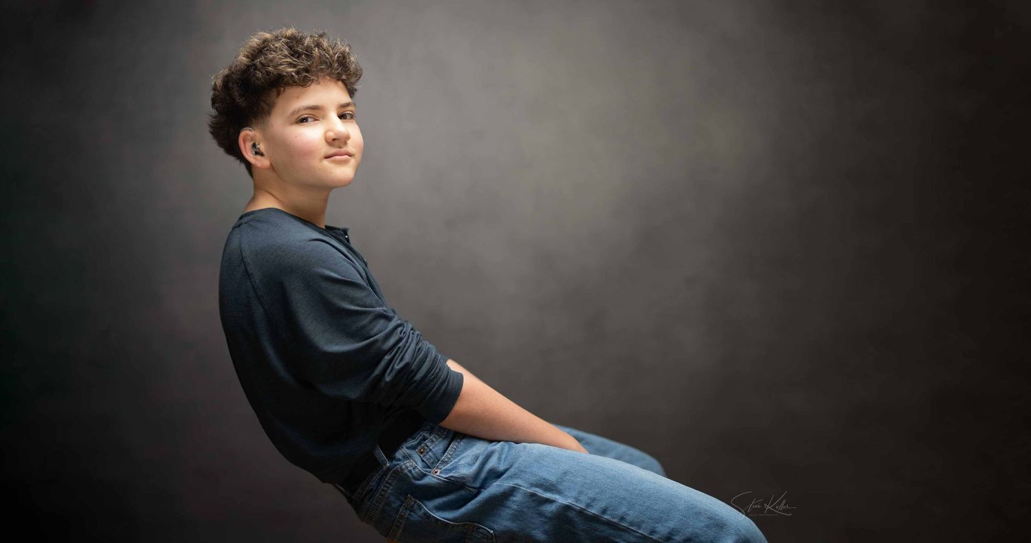 Young person with curly dark hair in a navy long-sleeve shirt and blue jeans sits against a gray studio backdrop in a portrait.