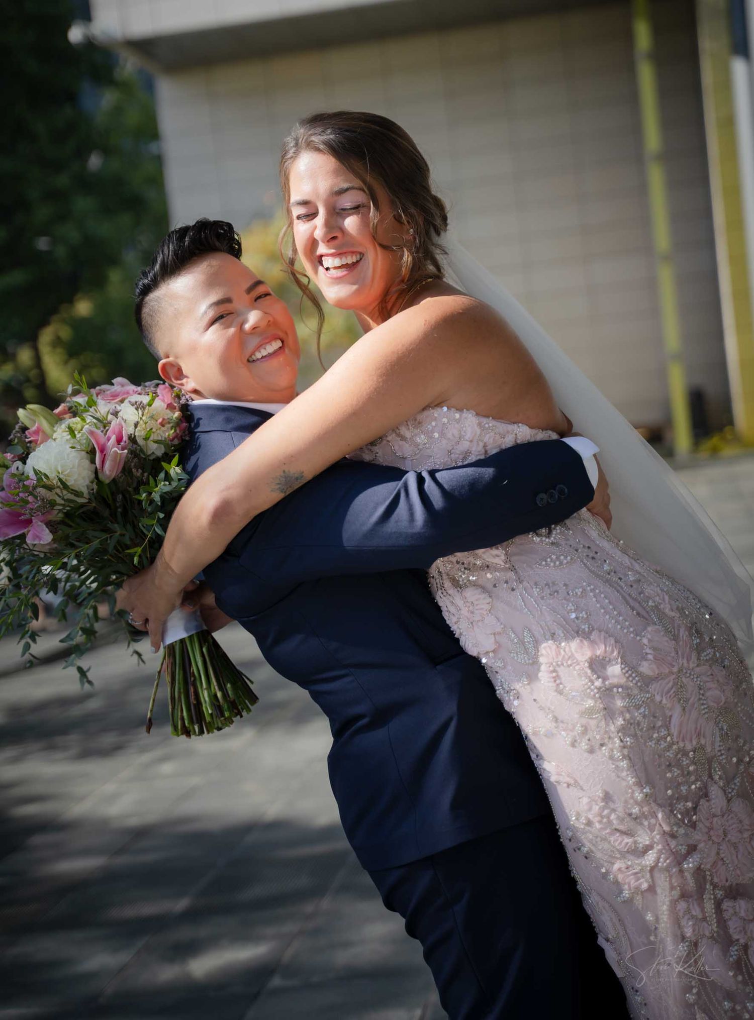 Bride in navy suit lifts her partner in a lace gown, both laughing while holding a pink floral bouquet.