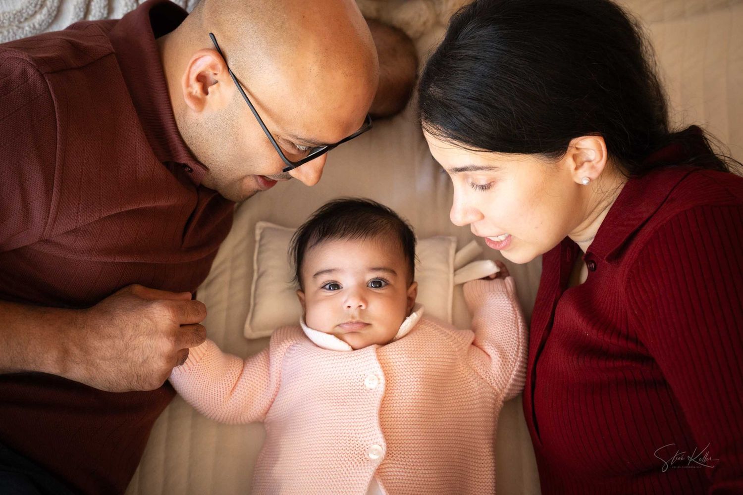 Parents lovingly gaze at their newborn baby lying between them on a soft bed in a tender family portrait.