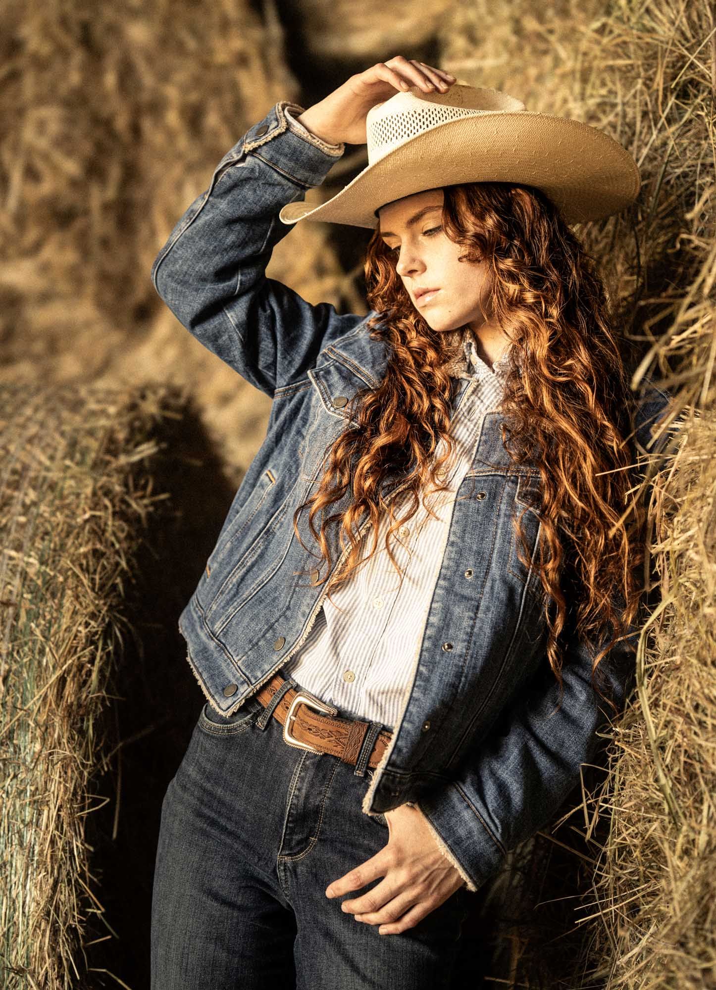 Woman in denim jacket, cowboy hat, and jeans leaning against hay bales with long curly auburn hair.