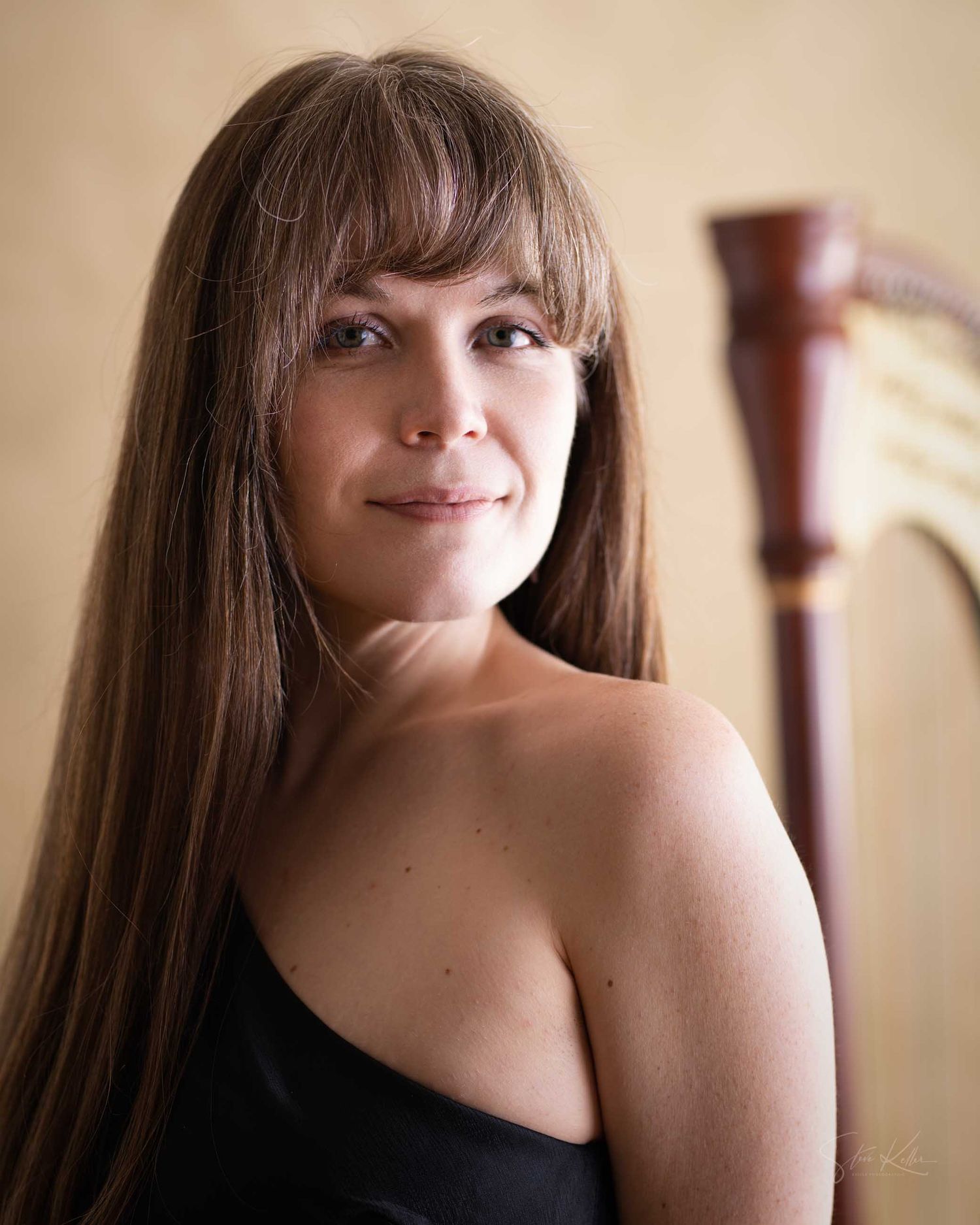 Woman with long brown hair smiling beside a harp, wearing a black strapless top in a professional portrait.