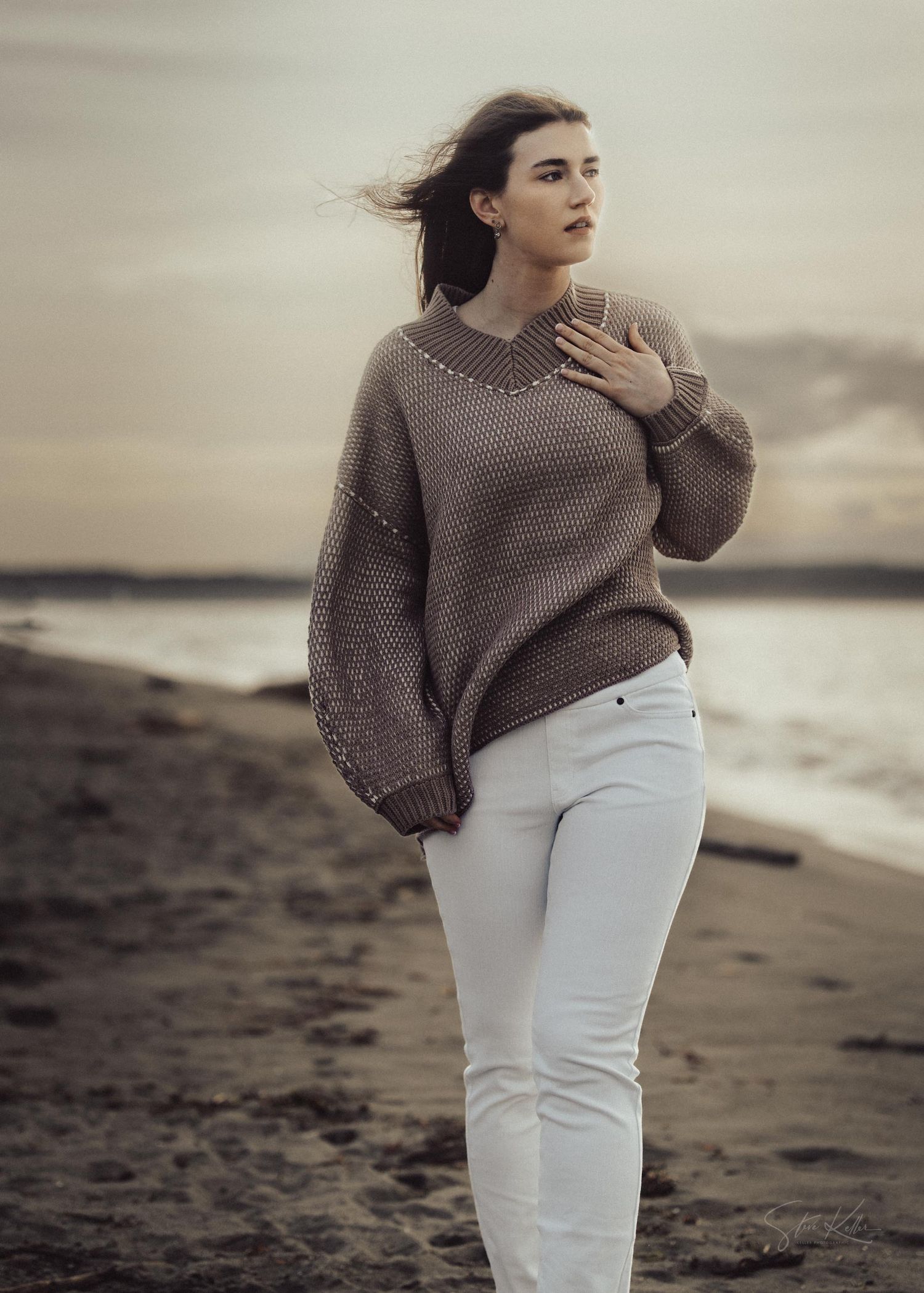 Person in oversized gray sweater and white pants poses dramatically on moody beach shoreline.