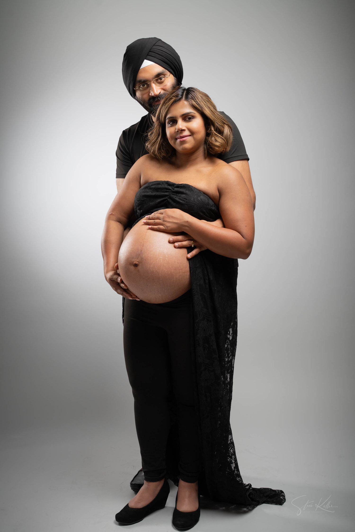 A pregnant woman in a black lace dress poses with her partner against a gray backdrop during a maternity photoshoot.