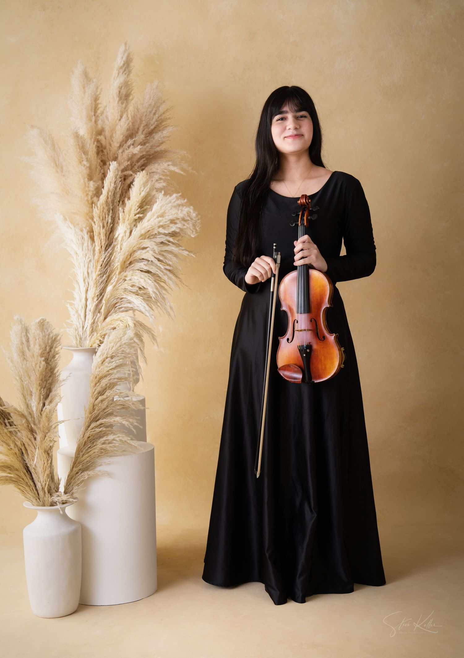 Musician in long black dress poses with violin against beige background with decorative pampas grass in white vases.
