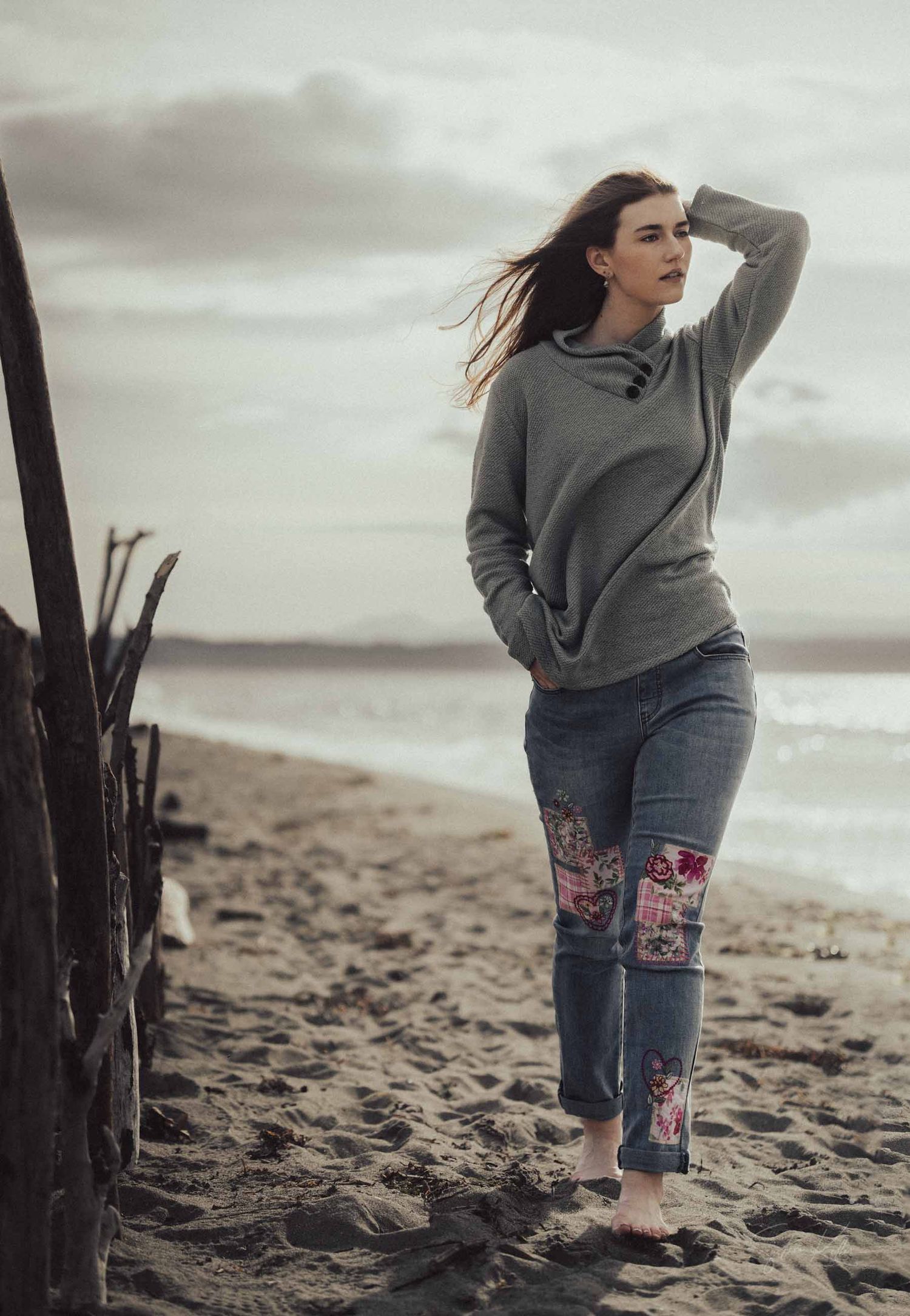 Person in grey sweater and patched jeans stands on sandy beach at sunset with moody cloudy sky in background.