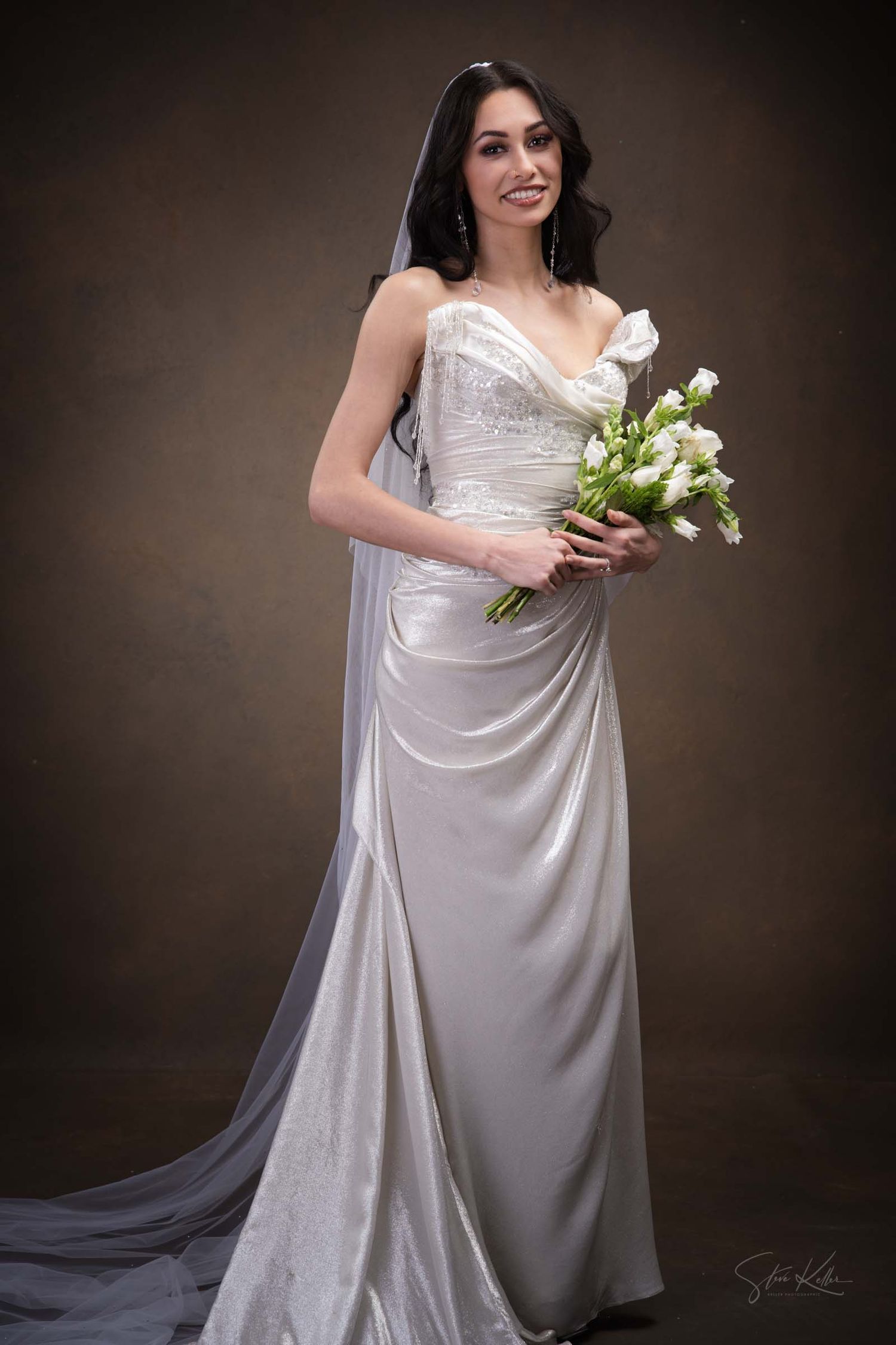 Elegant bride in flowing white strapless gown holds bouquet of white tulips against dark studio backdrop.