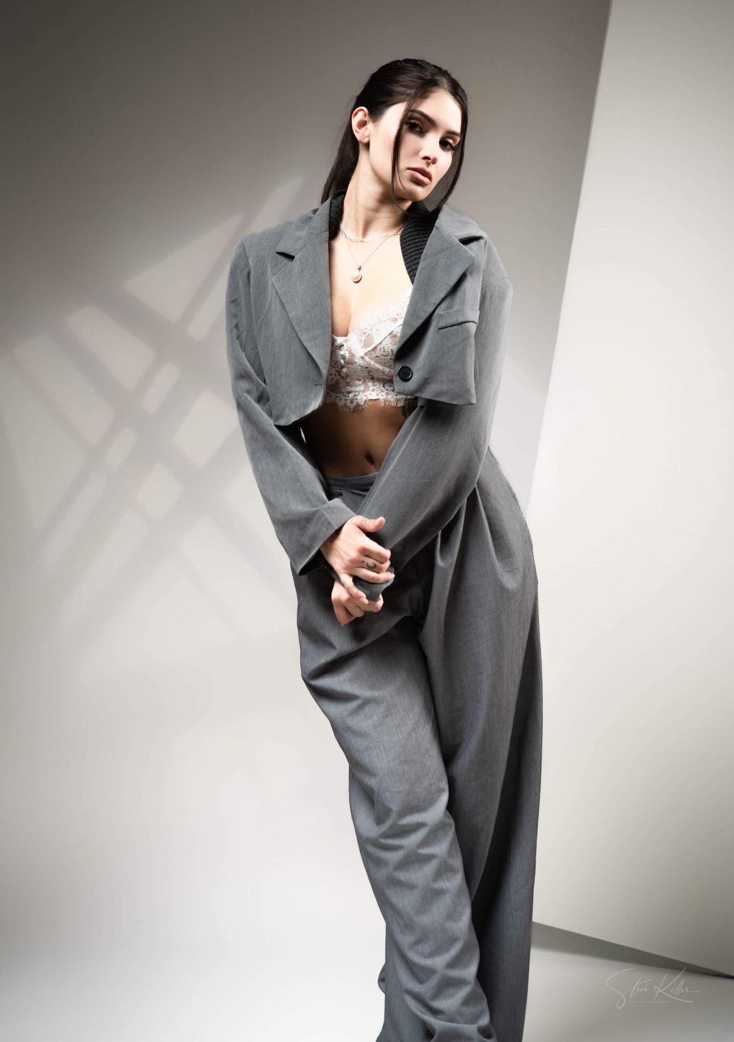 Fashion model in oversized grey suit and lace top poses dramatically against white wall with strong shadow effect.