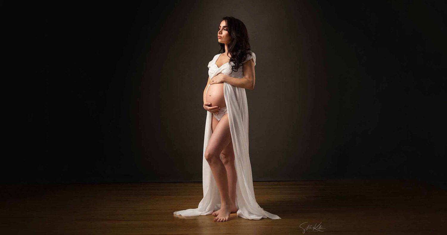 Pregnant woman in a flowing white sheer gown cradles her baby bump in a dark studio maternity portrait.