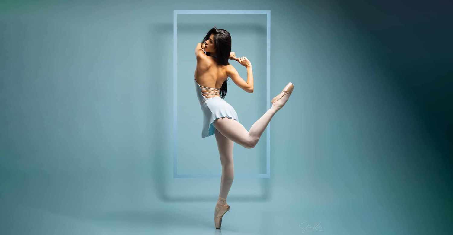 Female ballet dancer in a pale leotard balances en pointe with one leg raised in front of a soft teal backdrop.